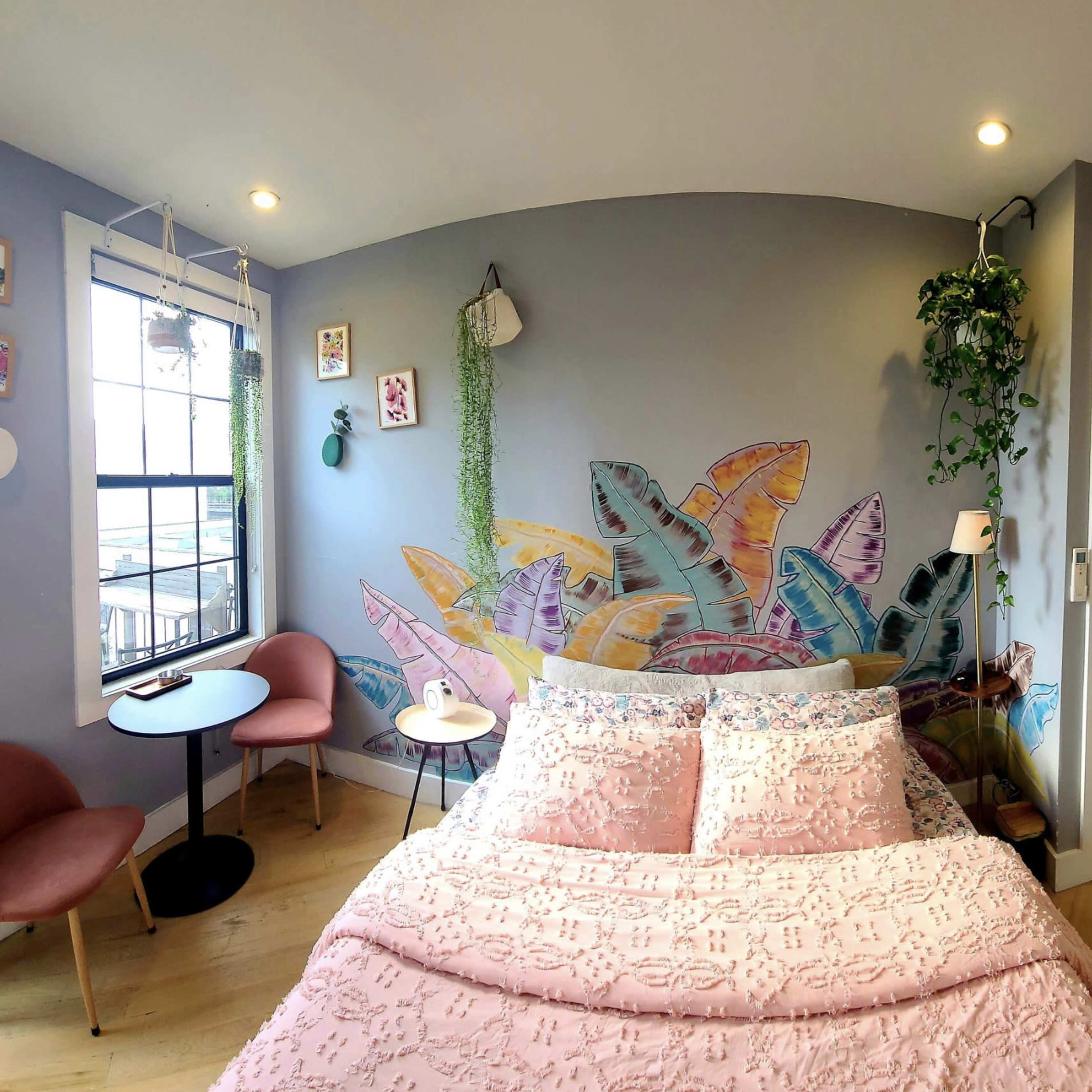 A cozy bedroom featuring a pastel-colored mural of fish on the wall, a pink bedspread, and two chairs with a small table nearby.