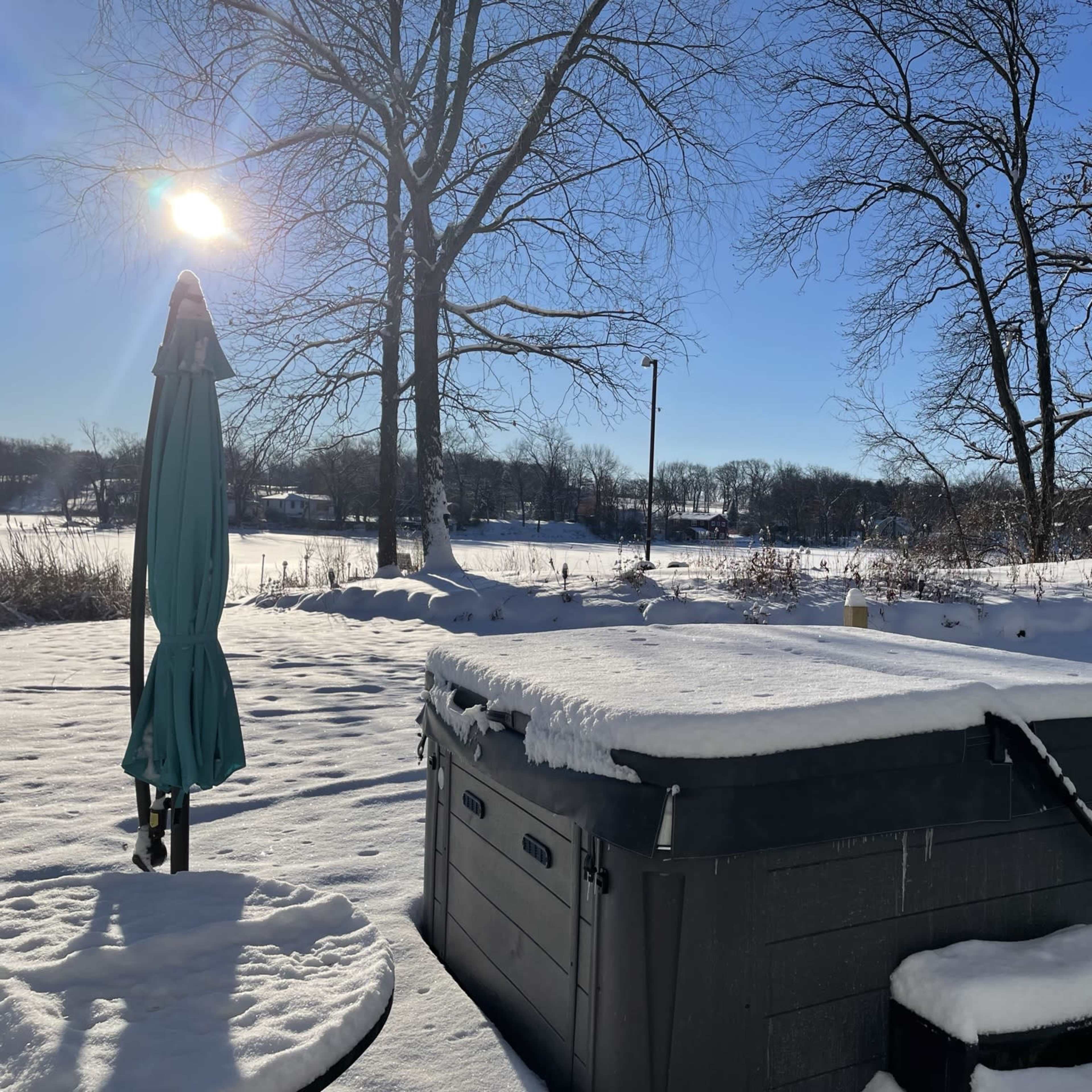A hot tub covered in snow sits in a backyard with bare trees and a clear blue sky.