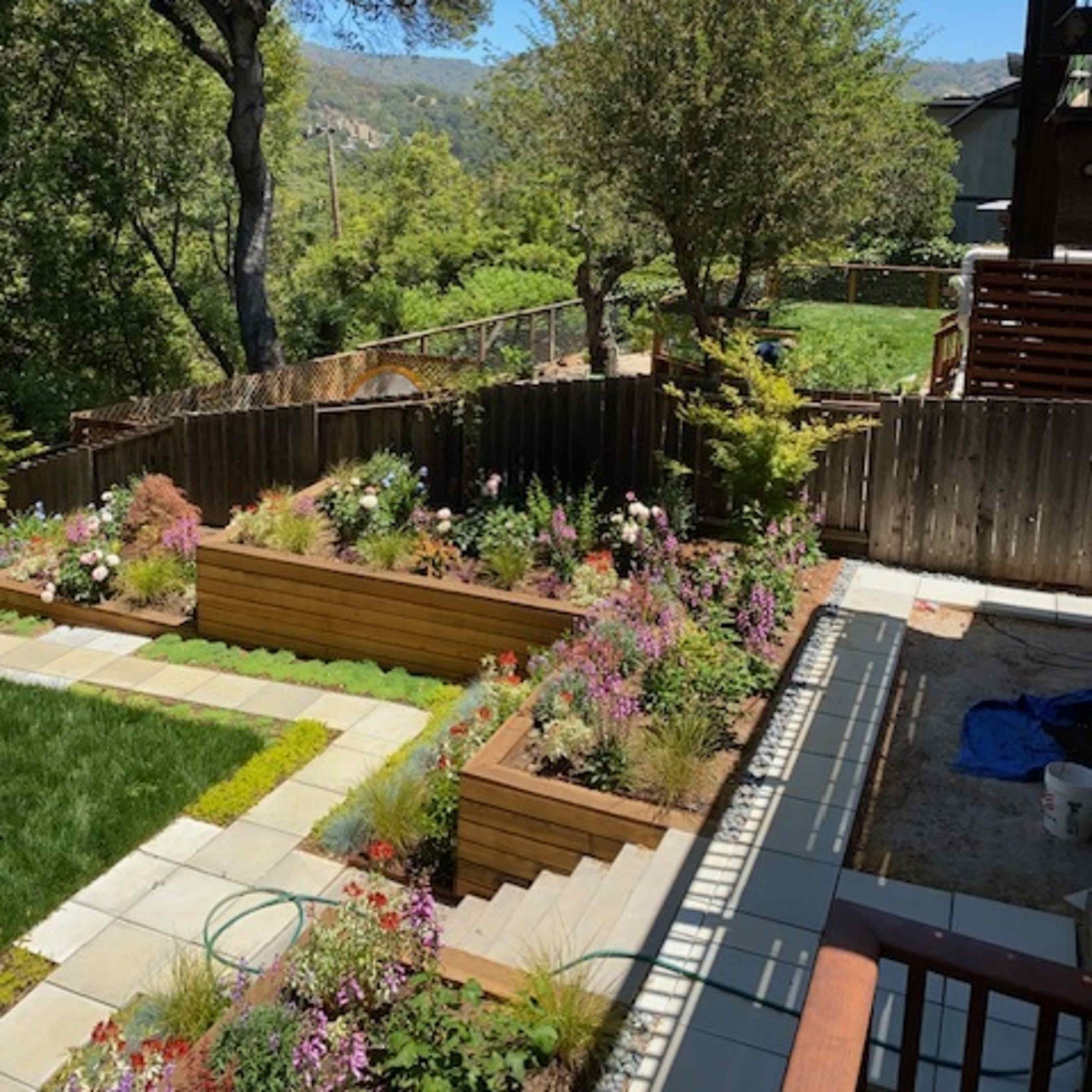 A landscaped backyard with raised garden beds filled with various plants and flowers, surrounded by trees and a scenic hill in the background.