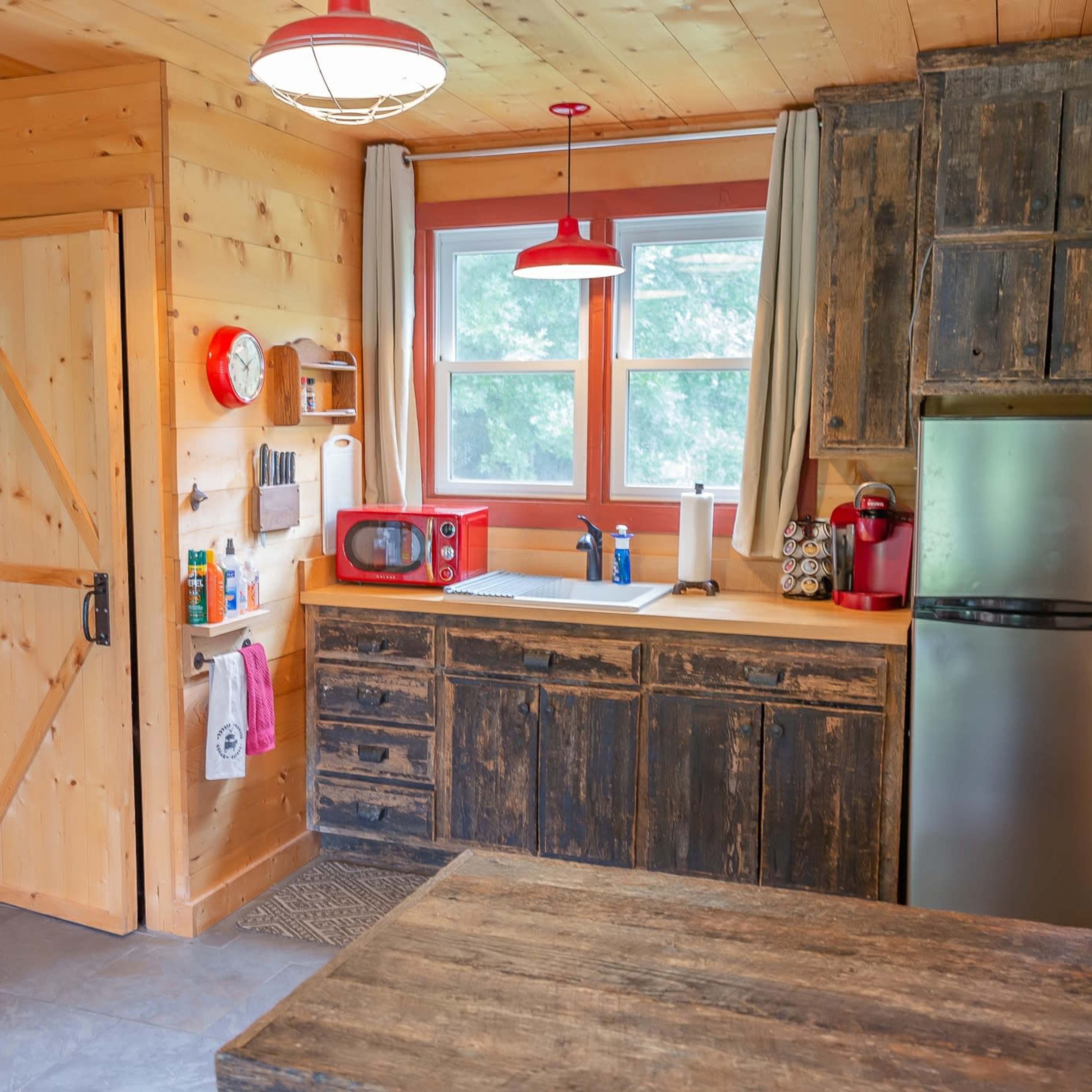 A rustic kitchen with wooden walls, a stone floor, a refrigerator, a red microwave, and a wooden table.