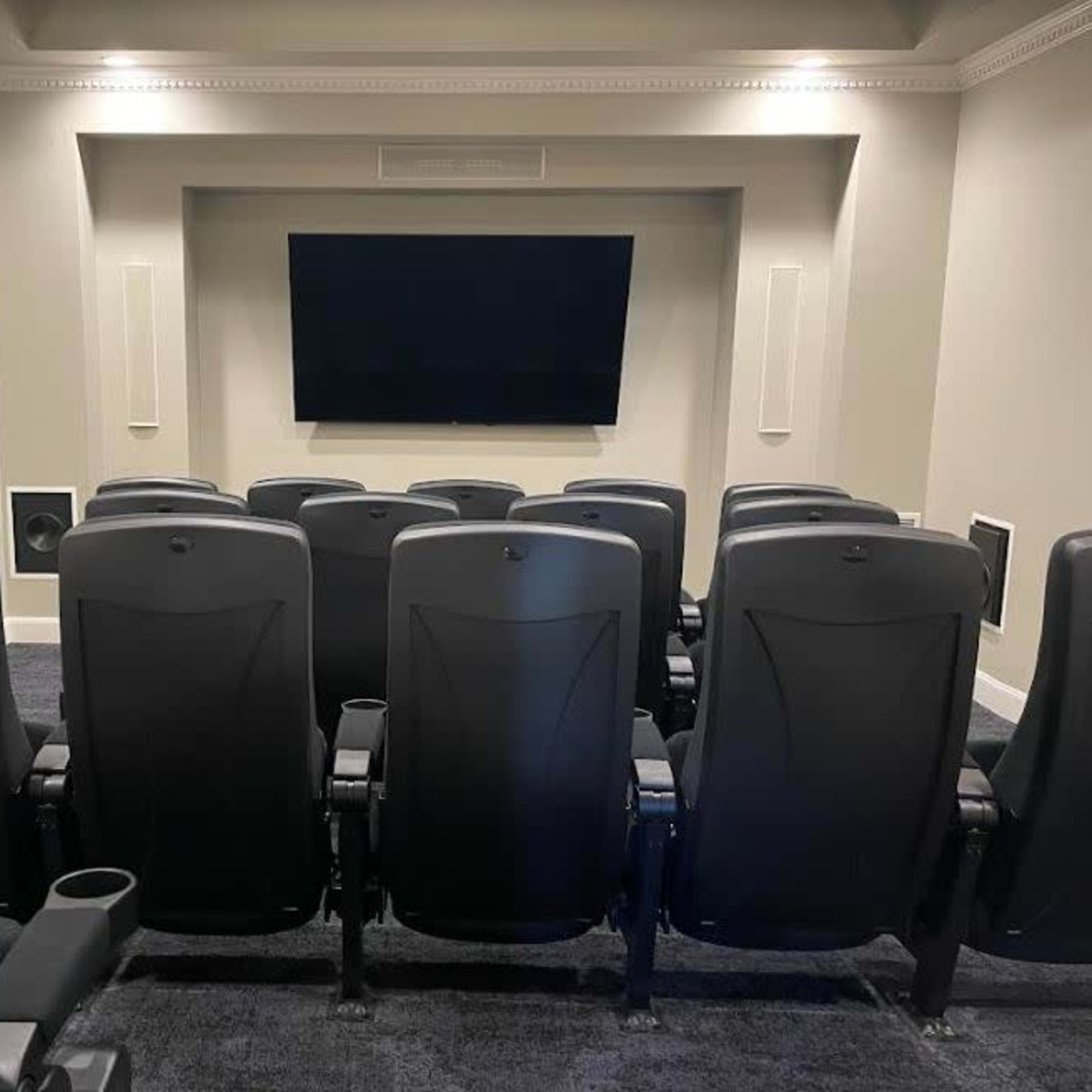 A home theater room with black reclining chairs arranged in rows facing a large flat-screen TV mounted on the wall.