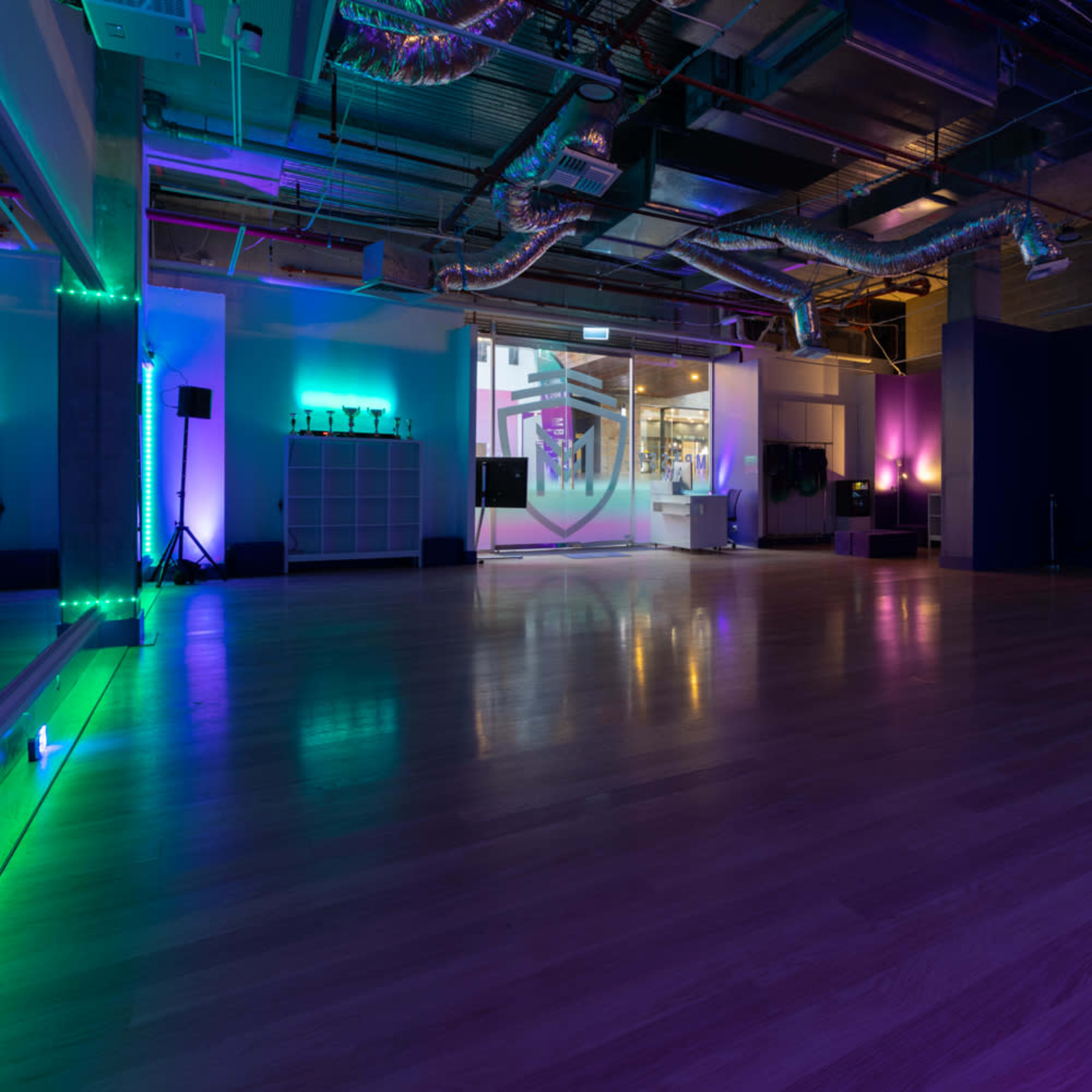 A spacious dance studio with a wooden floor, dim lighting, and a prominent wall featuring the name "The M Ambassador Dance."