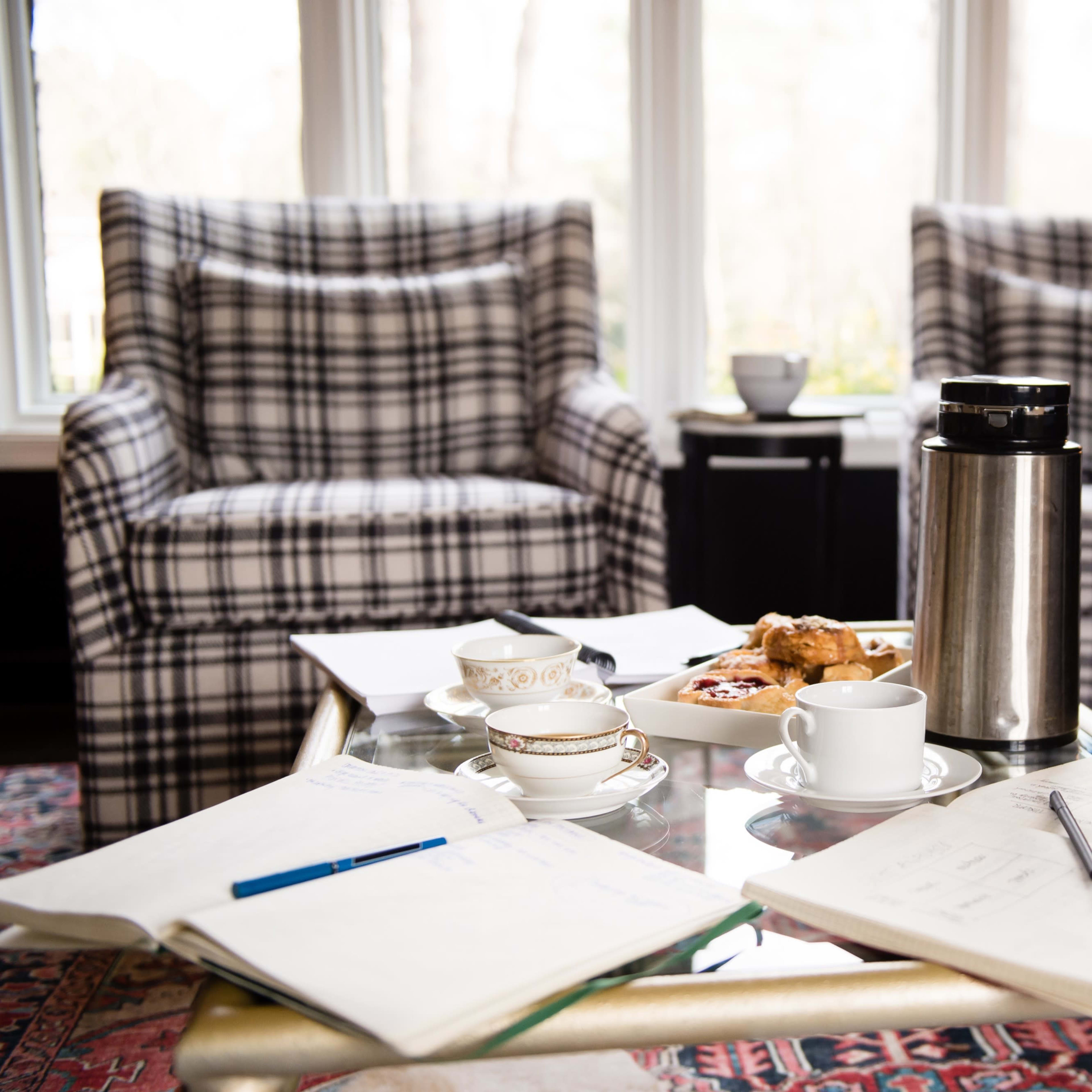 A cozy seating area features two plaid armchairs, a coffee thermos, pastries on a plate, and scattered notebooks on a glass coffee table.