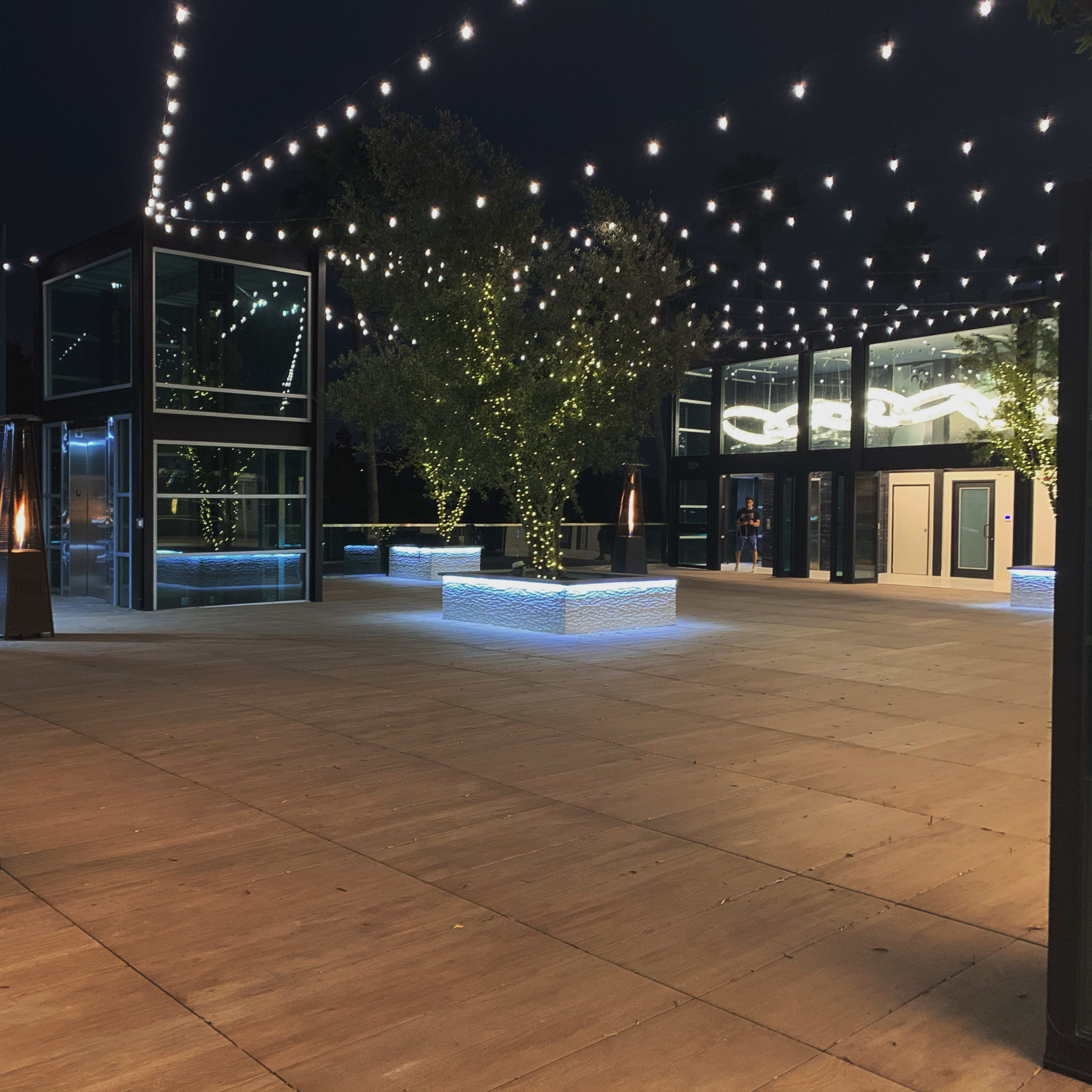 An outdoor venue at night, featuring a wooden floor, illuminated trees, string lights overhead, and modern glass structures.