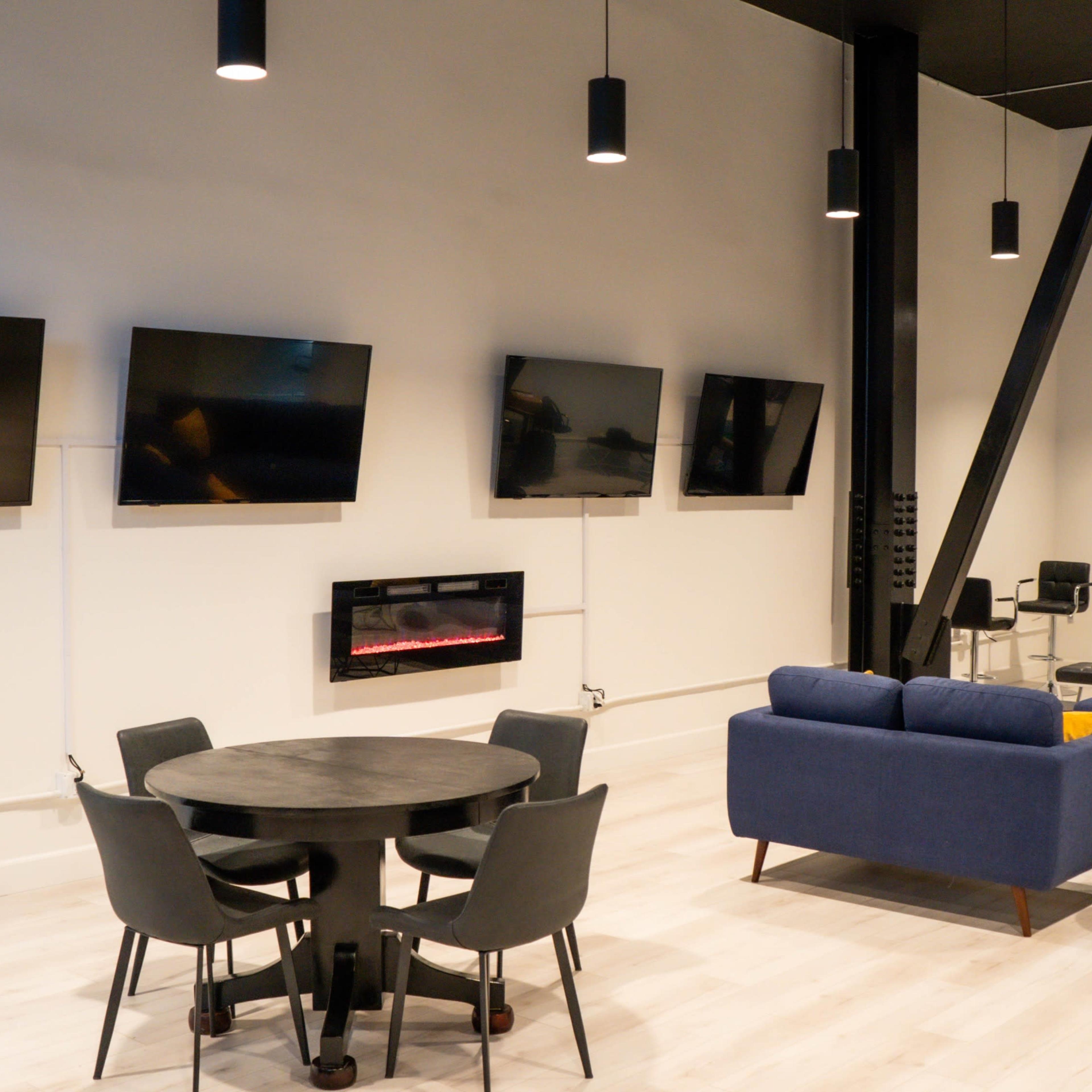 A modern living area with a circular dining table and chairs, a blue sectional sofa, a wall-mounted electric fireplace, and several flat-screen televisions mounted on the wall.