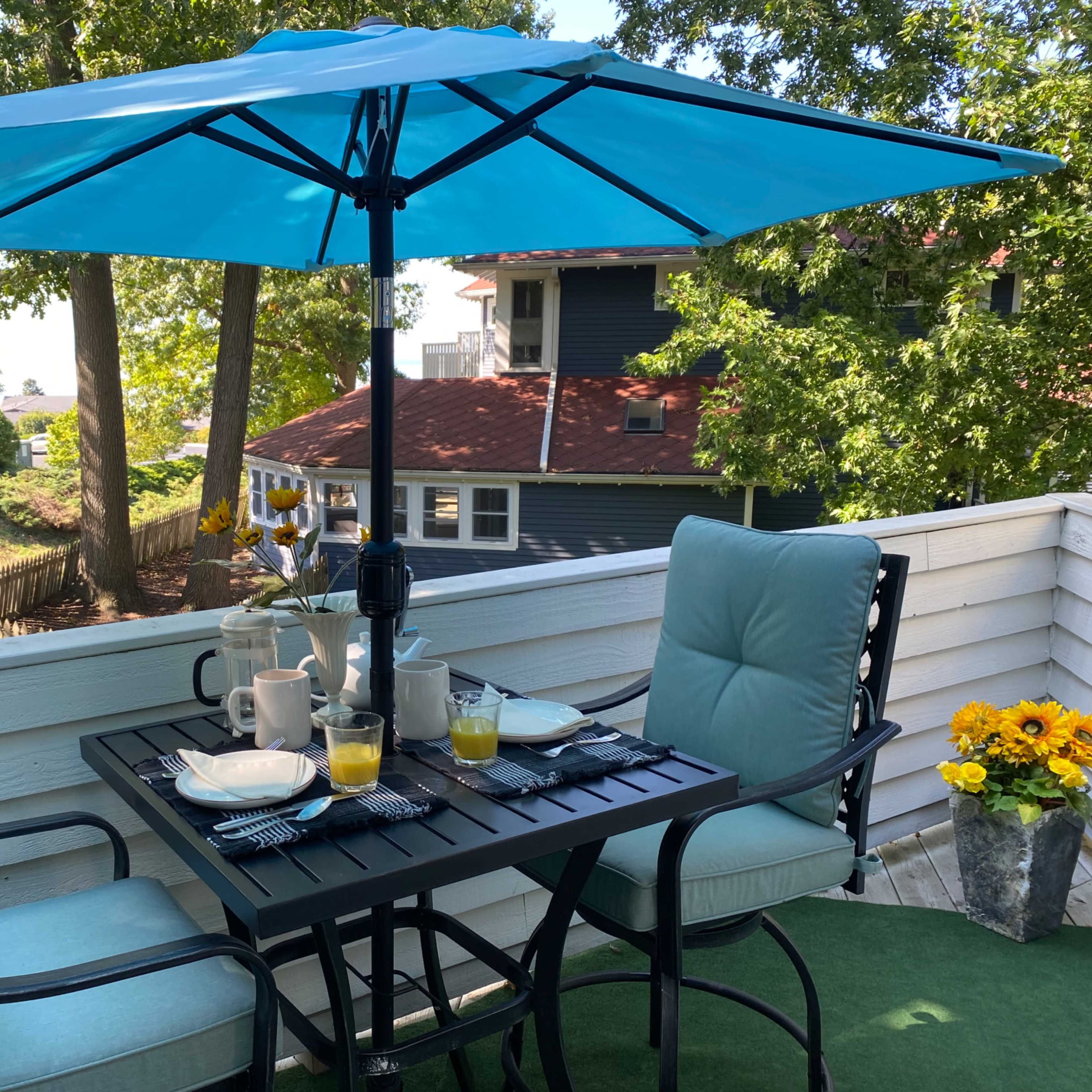 A table set for breakfast is positioned under a turquoise umbrella on a balcony surrounded by trees.