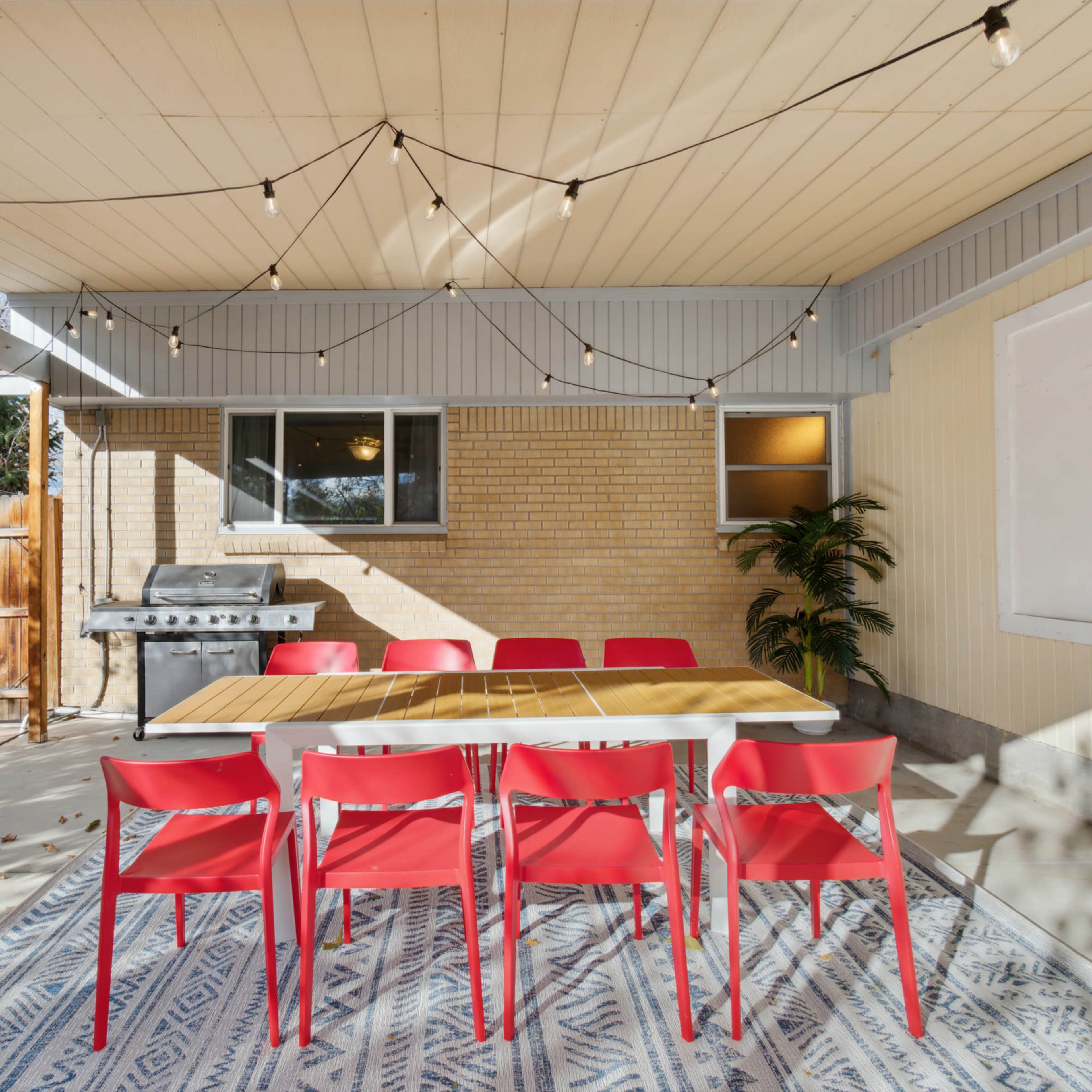 An outdoor patio area with a long wooden table surrounded by six red chairs, string lights overhead, and a barbecue grill in the corner.