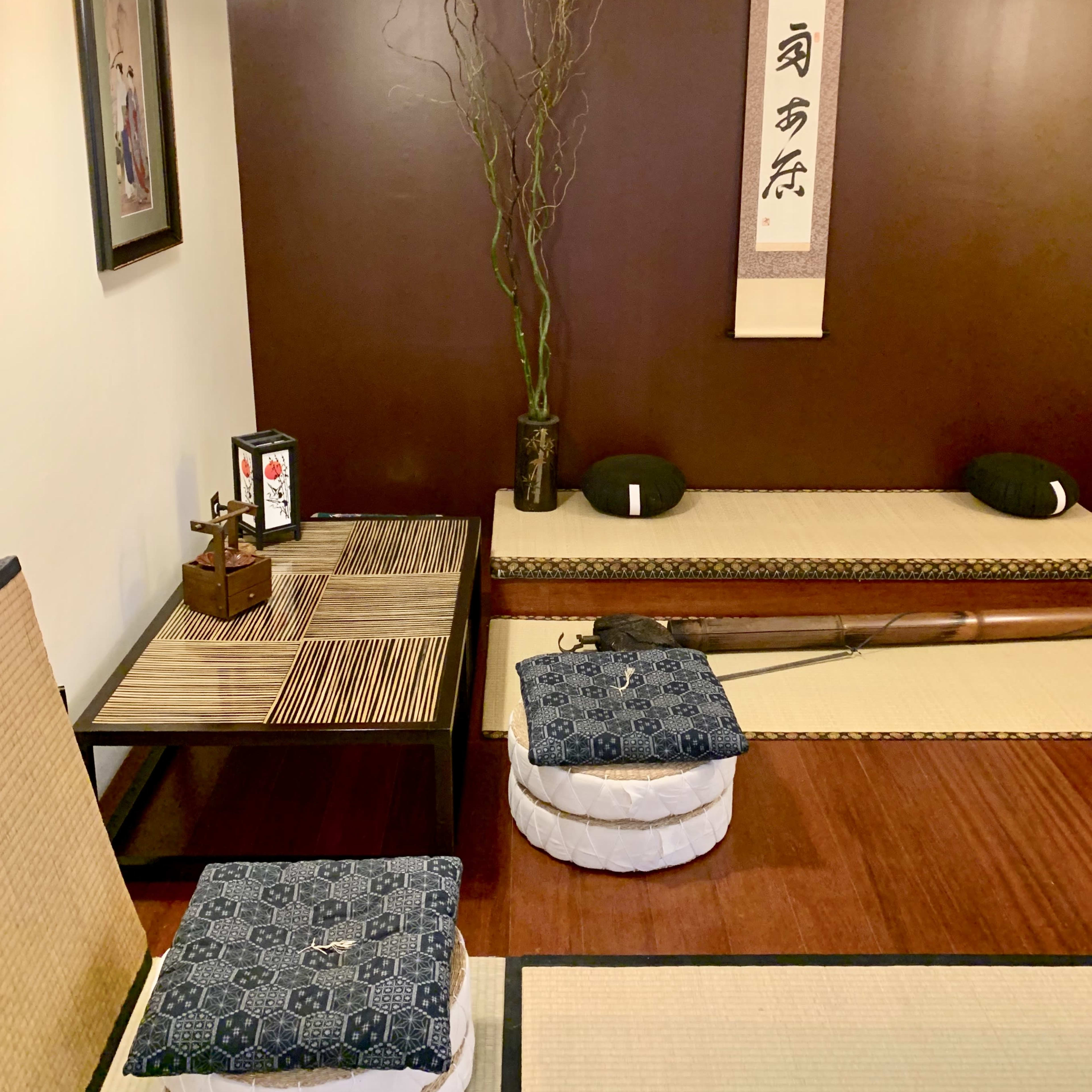 A traditional Japanese-style room with tatami flooring, low seating, and decorative elements including a hanging scroll and a plant.