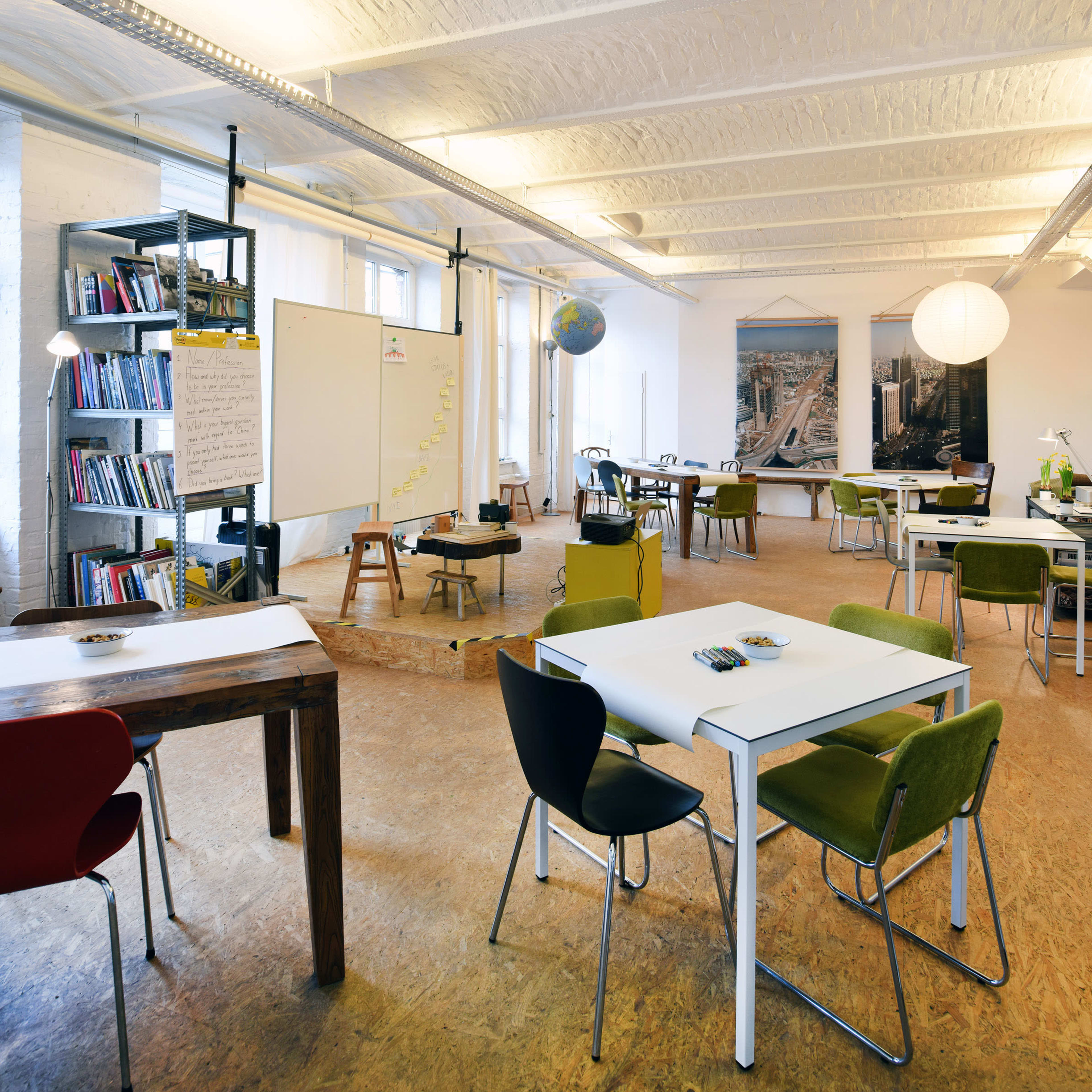 A modern, open office space with several tables and chairs, bookshelves, a whiteboard, and decorative elements like a globe and wall art.