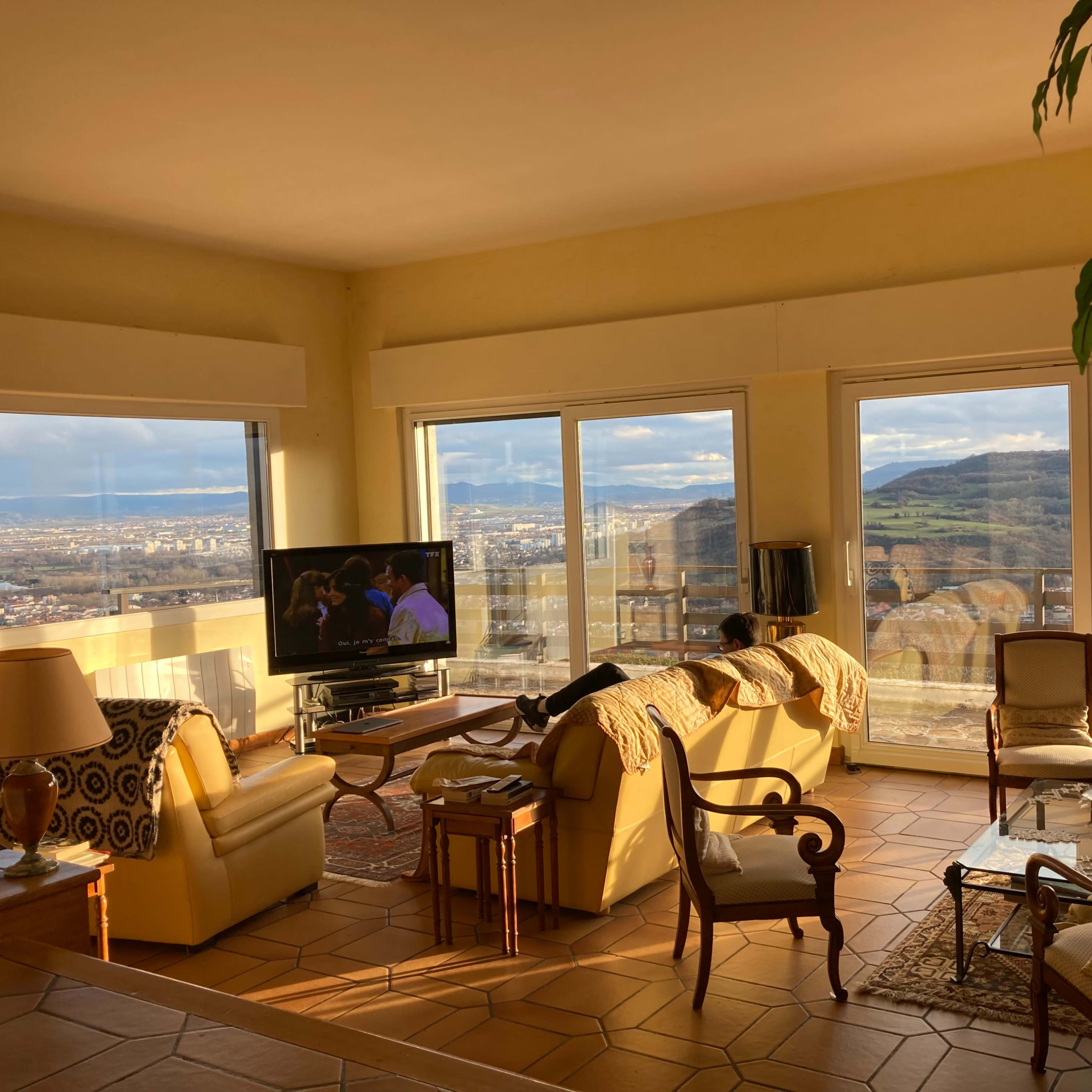 A living room with large windows overlooking a cityscape, featuring beige furniture, a television, and a plant.