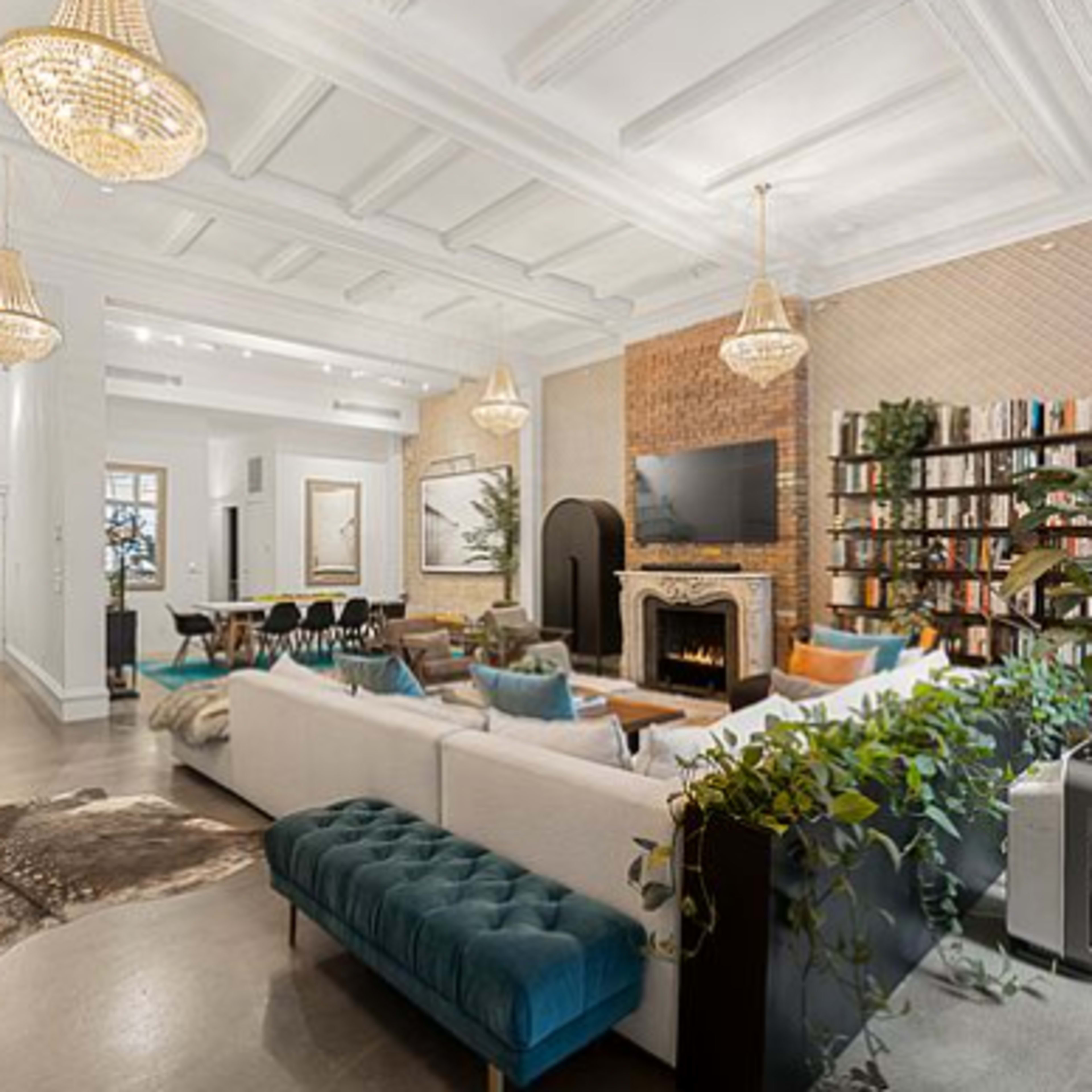 A spacious living room featuring a white sectional sofa, a blue velvet bench, a fireplace, and a large bookshelf filled with books, all illuminated by pendant chandeliers.