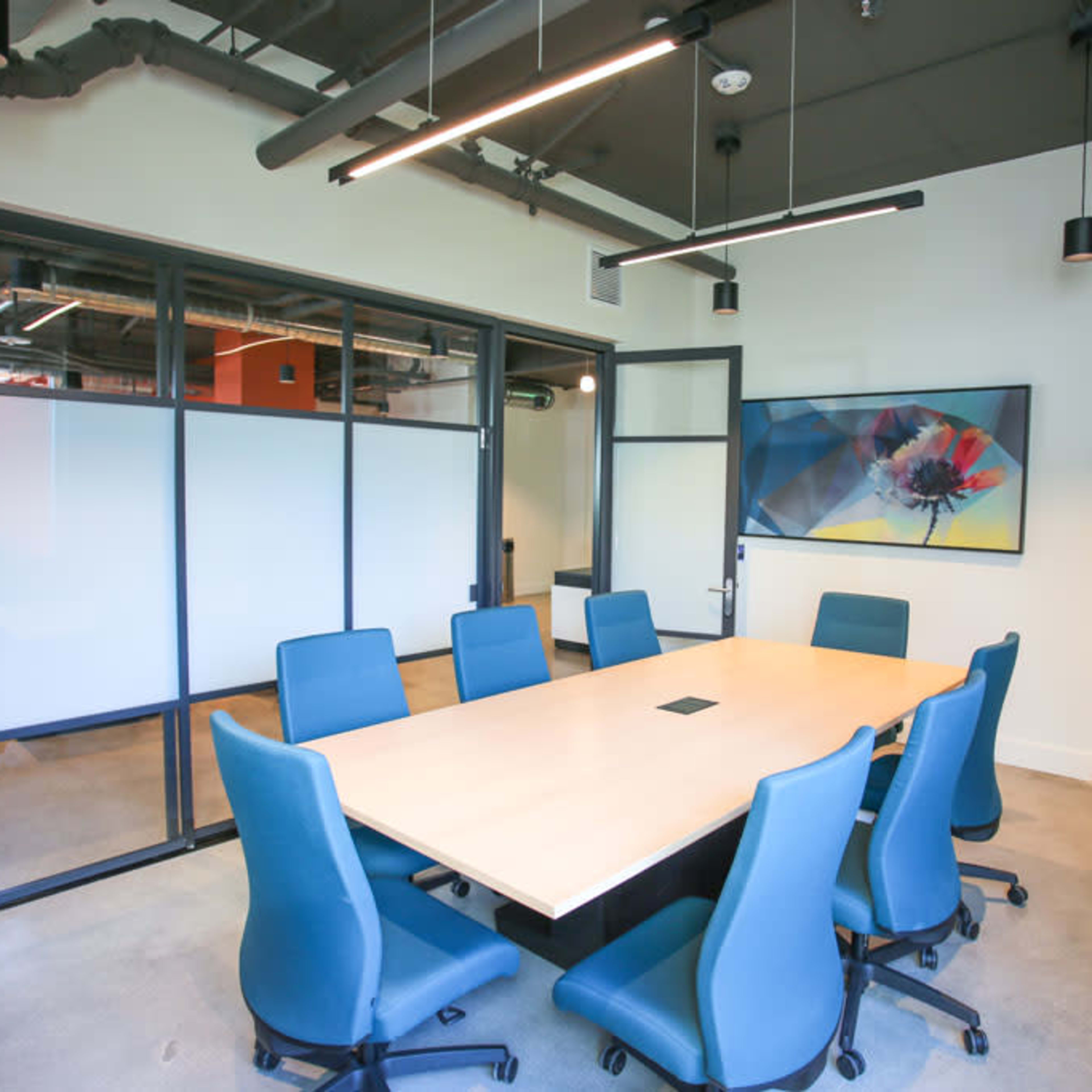 A modern conference room featuring a large rectangular table surrounded by blue chairs, with large windows and a potted plant.