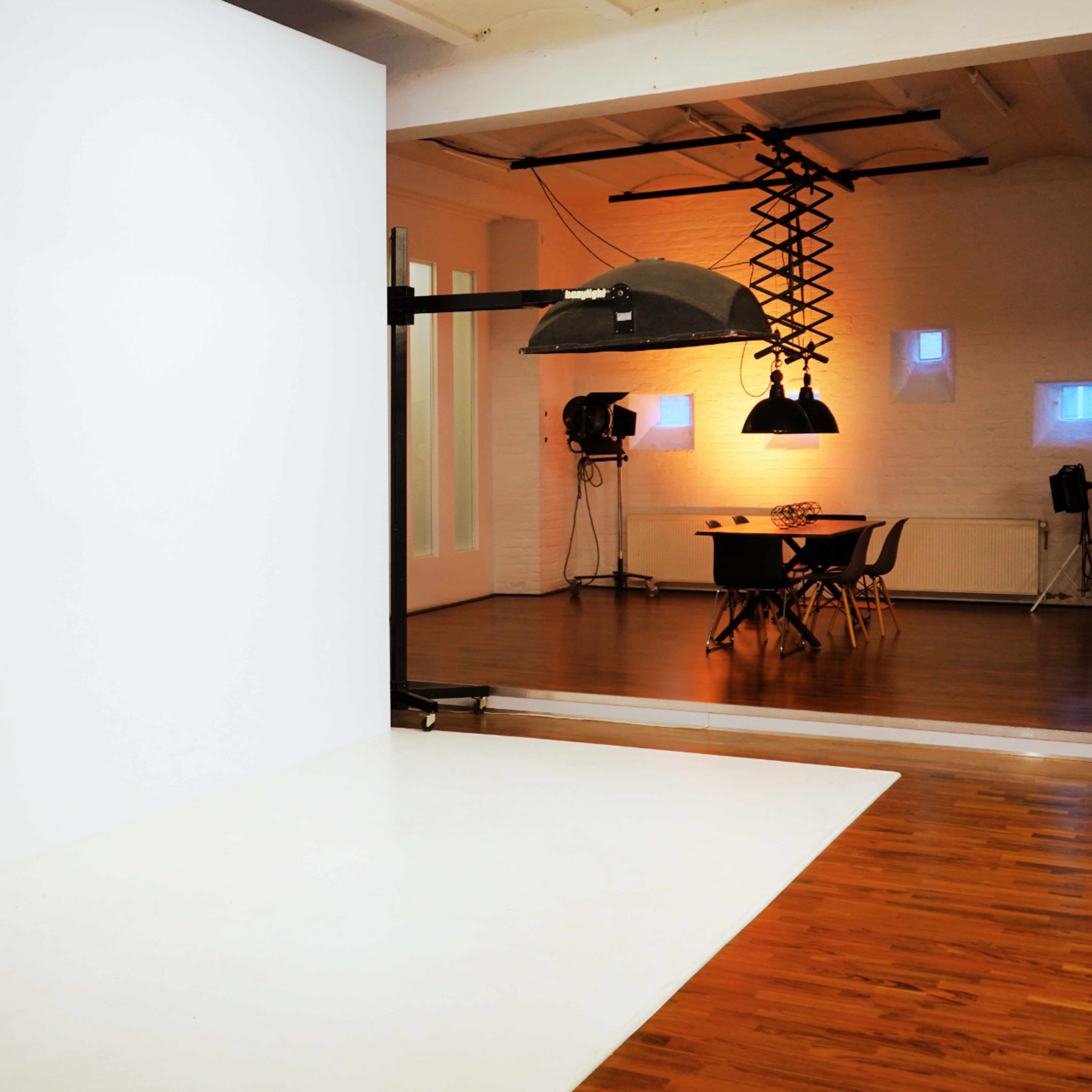 A photography studio featuring a white backdrop, a large light fixture, and a table with chairs in a well-lit space.