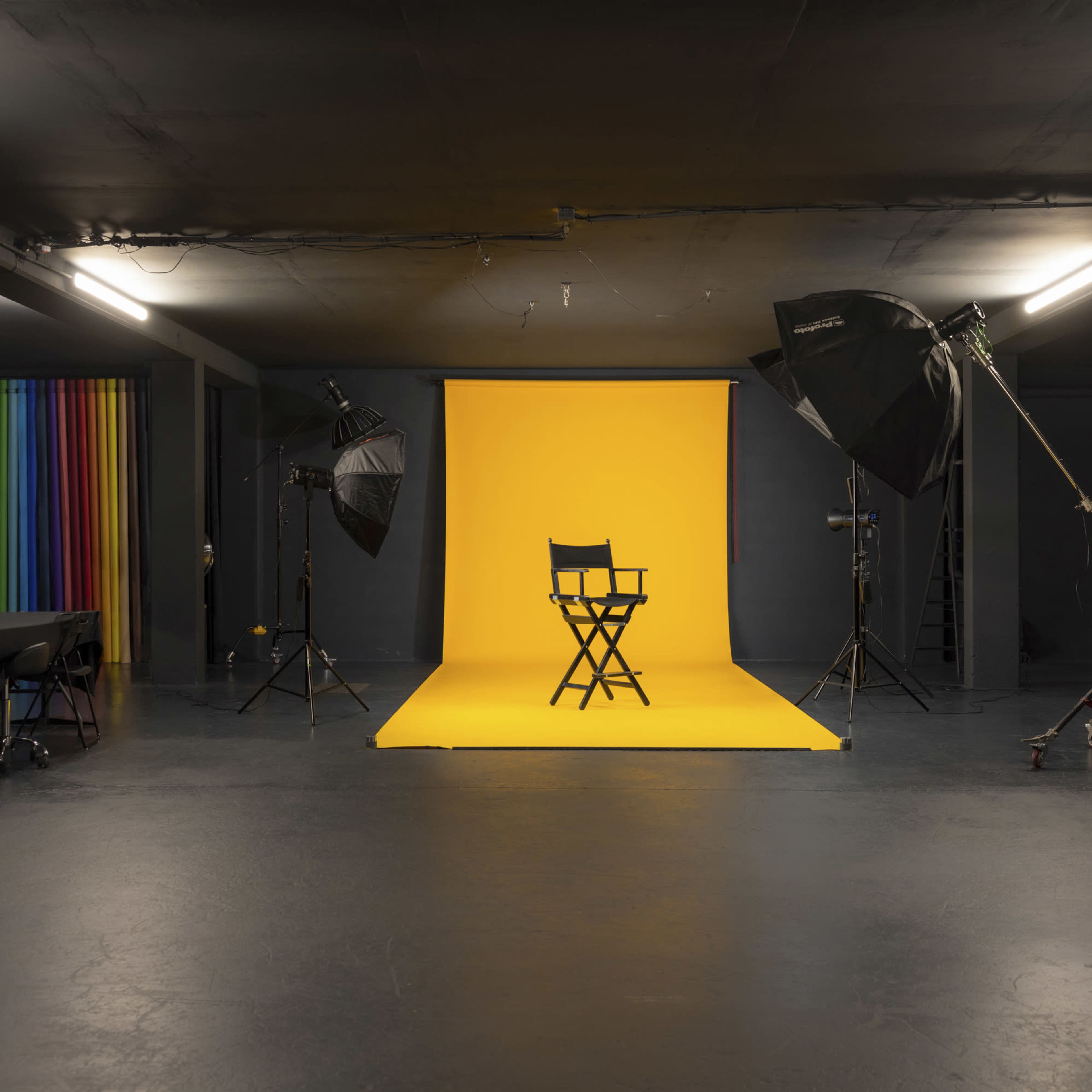 A photography studio features a black floor and walls, with a yellow backdrop, a director's chair at the center, and various lighting equipment positioned around.