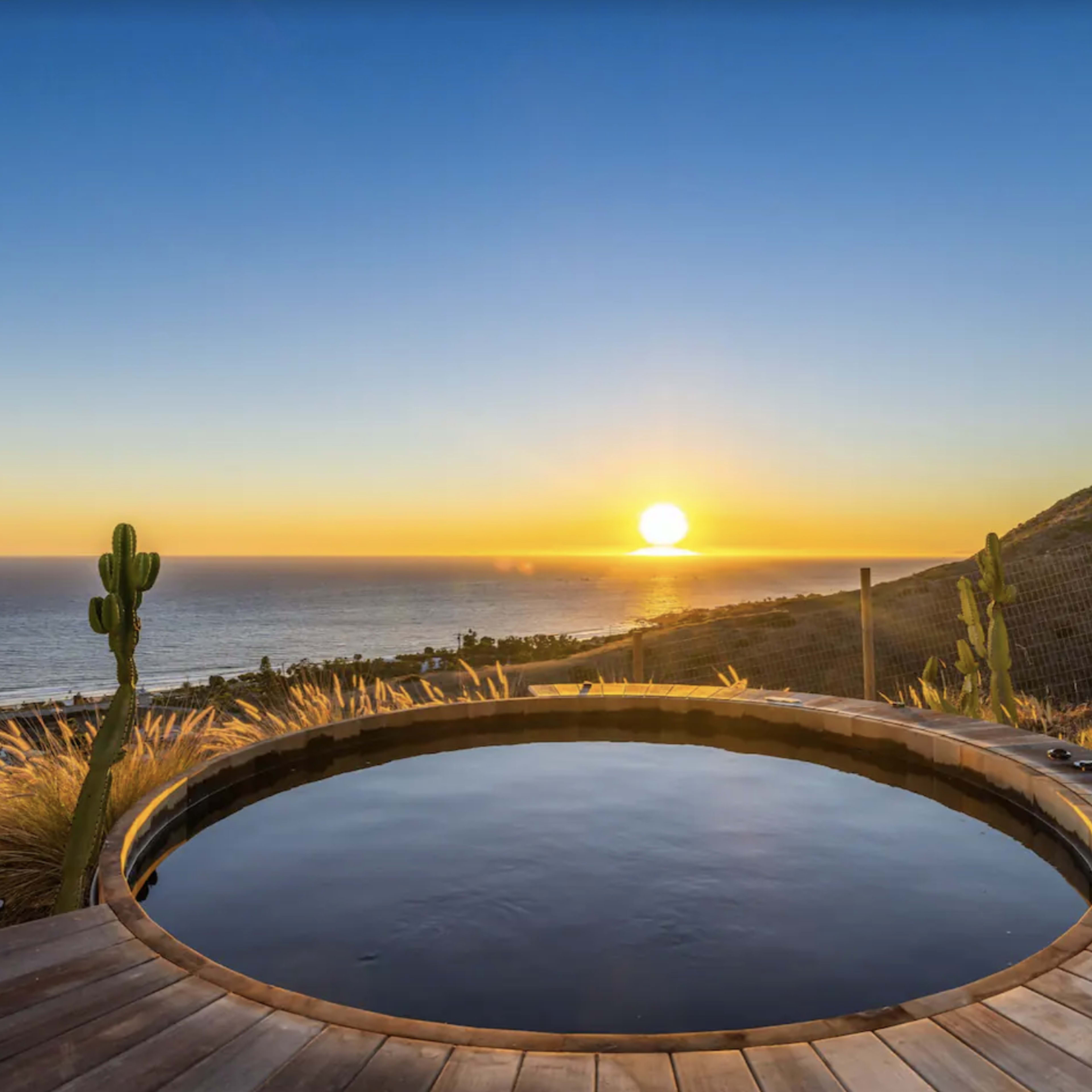 A circular wooden hot tub overlooks the ocean at sunset, framed by desert vegetation.