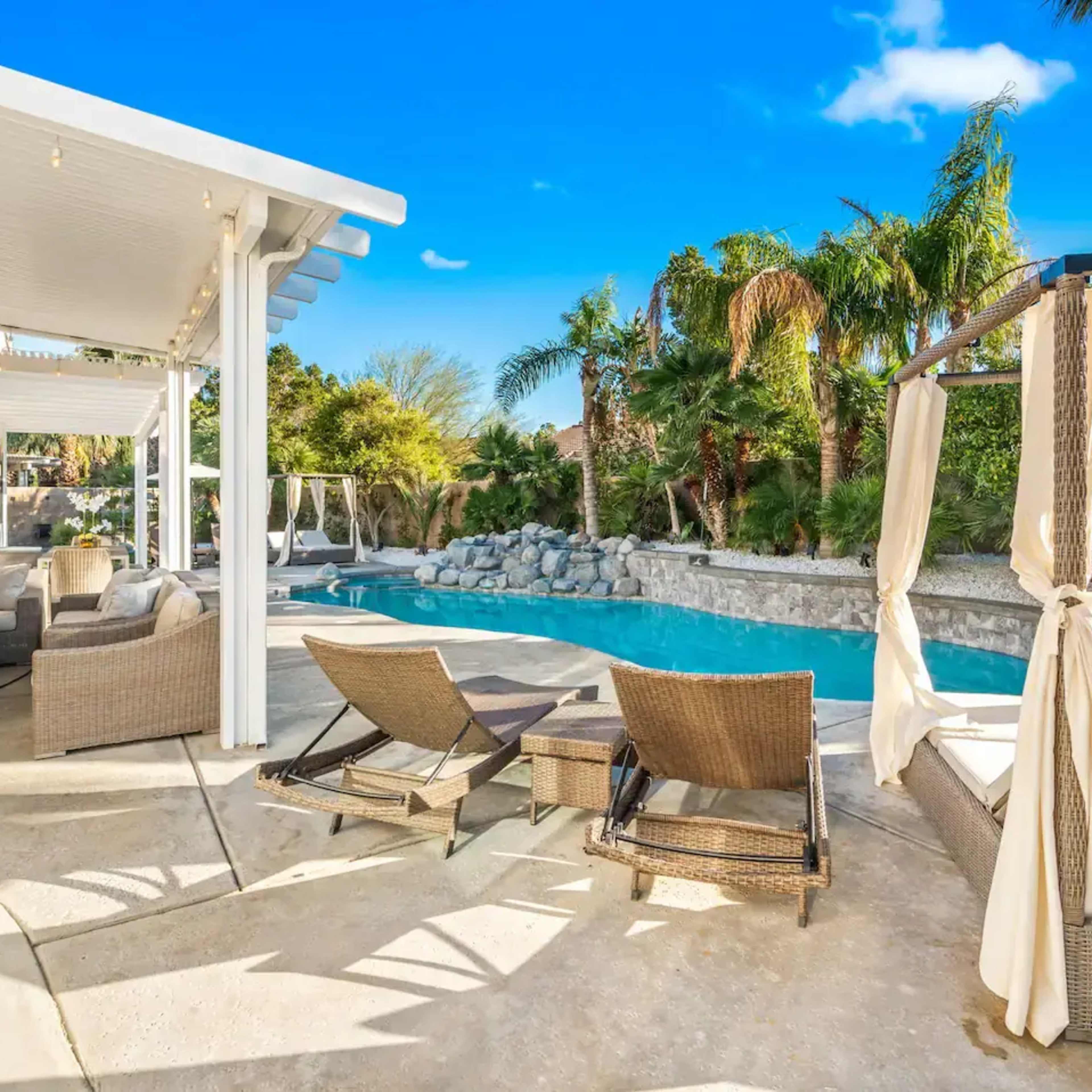 A backyard space with a swimming pool, lounge chairs, and a covered patio area surrounded by palm trees and landscaping.
