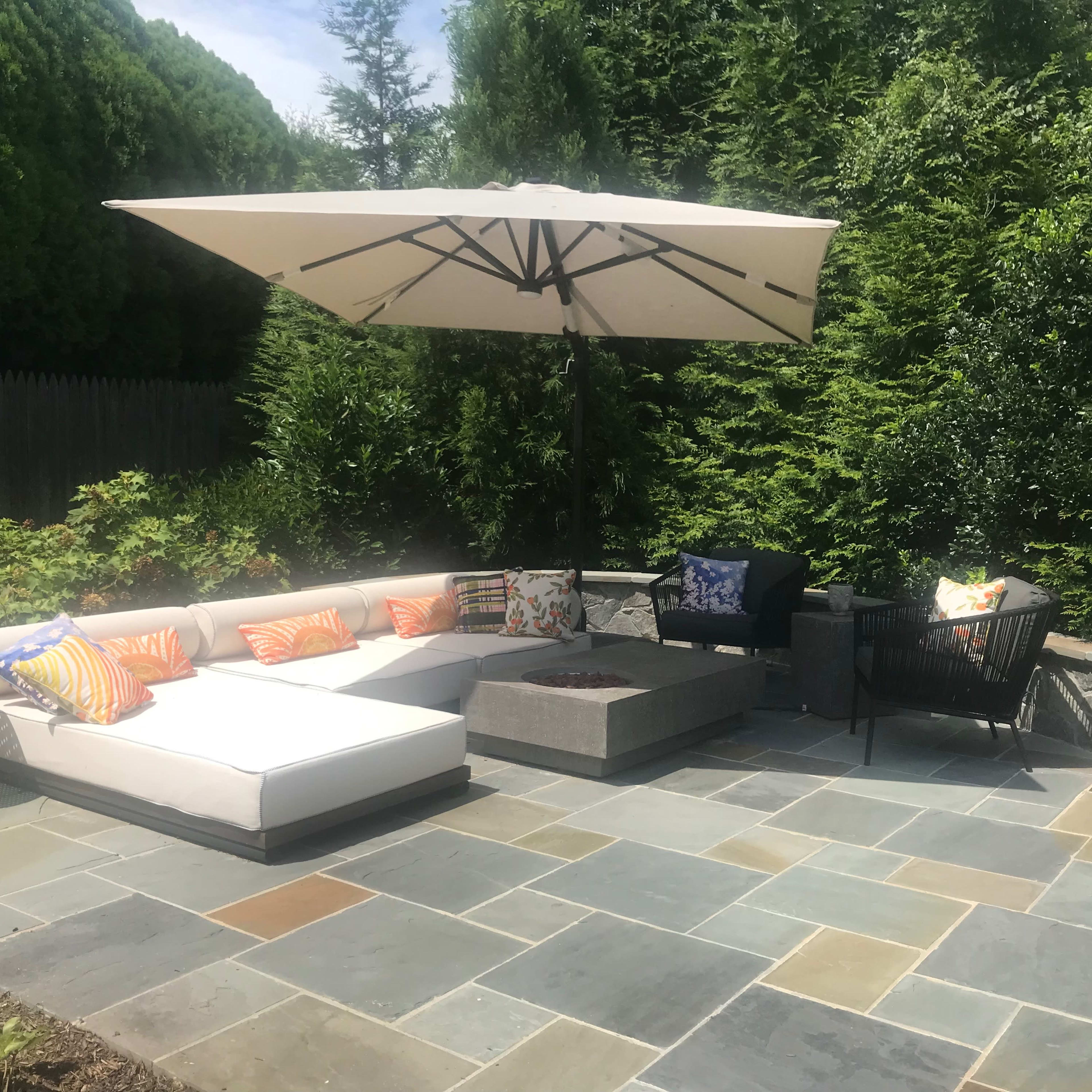 A patio area features a sectional sofa with colorful pillows, a central coffee table, and a large umbrella, surrounded by greenery and stone flooring.