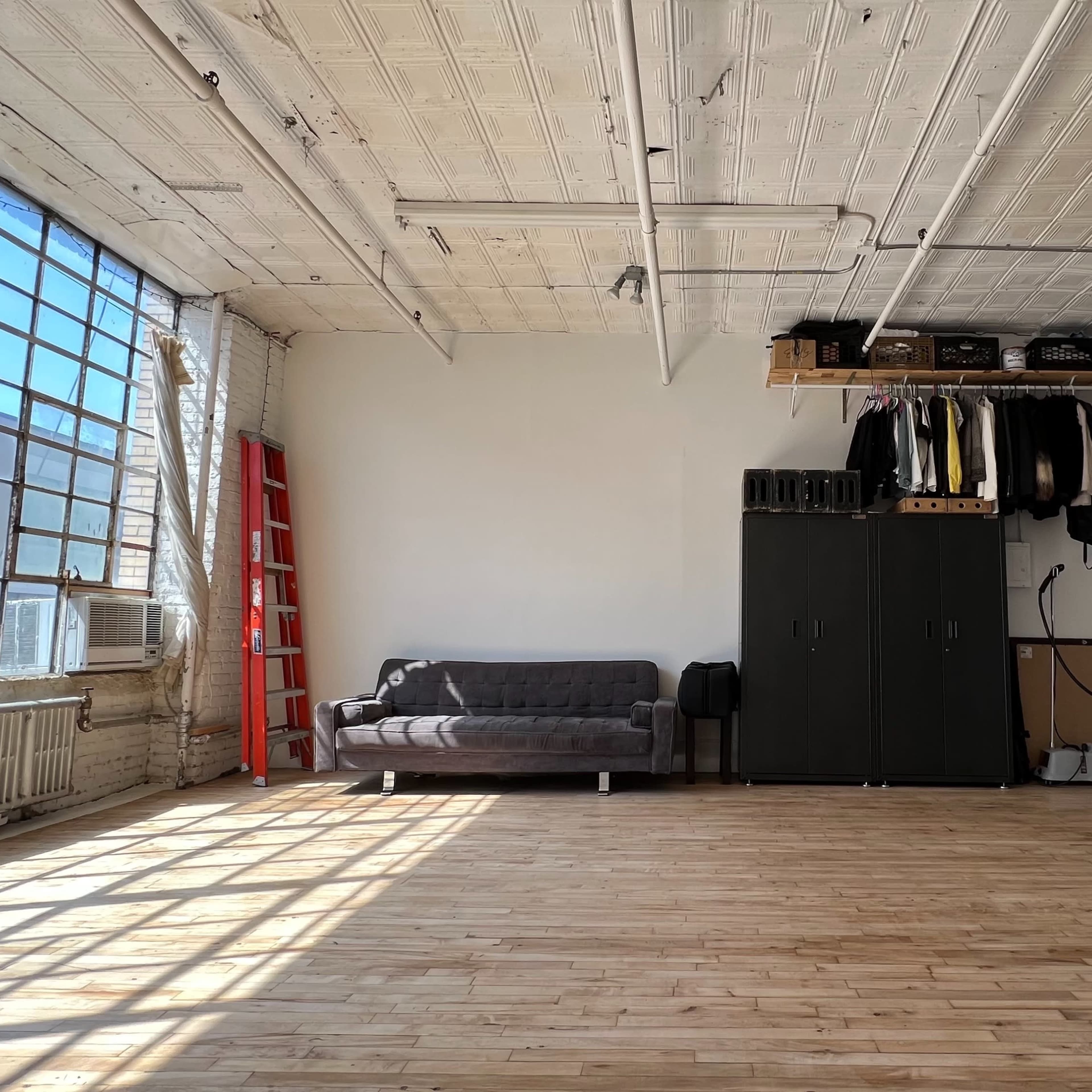 A spacious studio with large windows, wooden flooring, and a lounge area featuring a couch, storage cabinets, and various studio equipment.