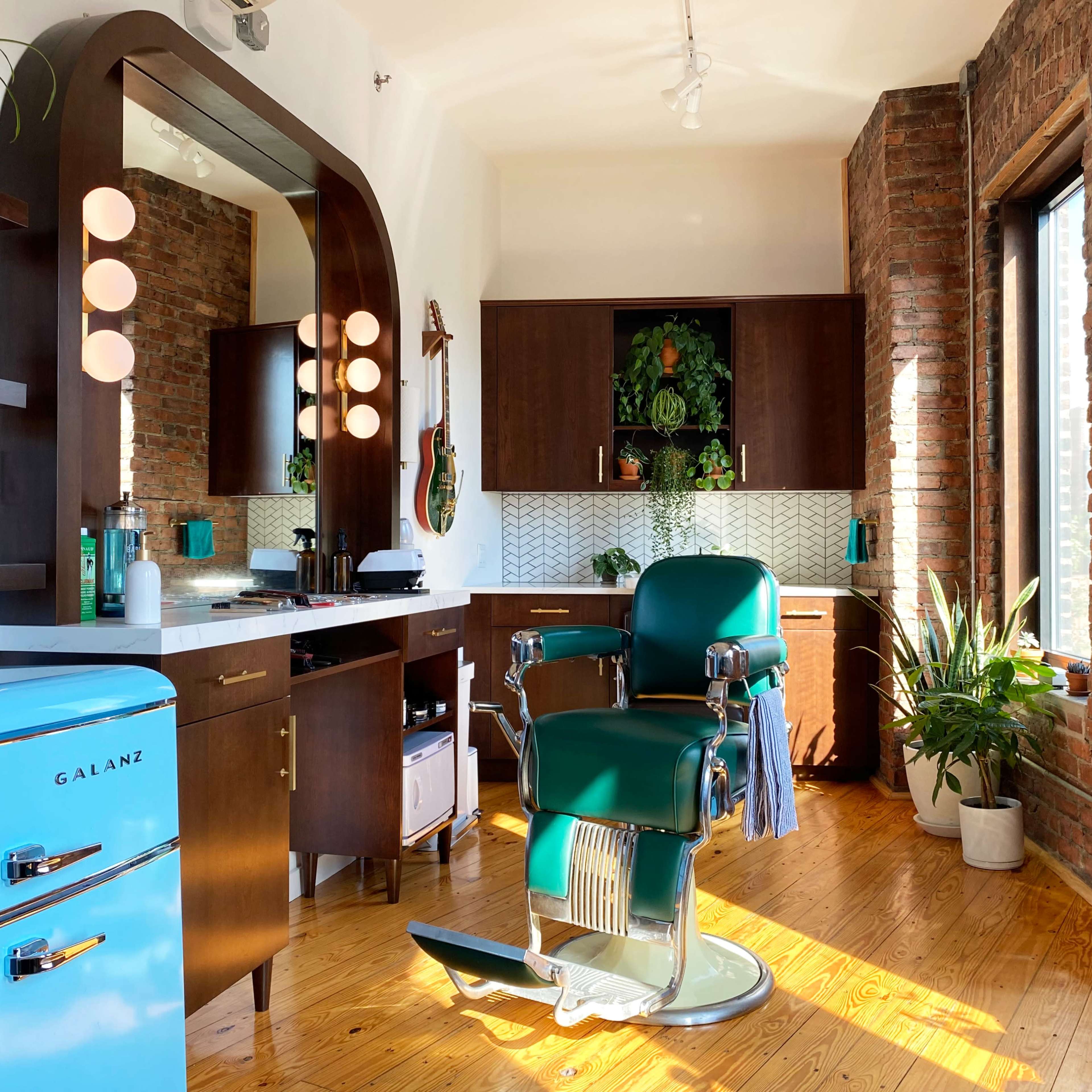 A bright barbershop interior featuring a vintage blue refrigerator, a green barber chair, and wooden cabinetry against a backdrop of exposed brick walls and plants.
