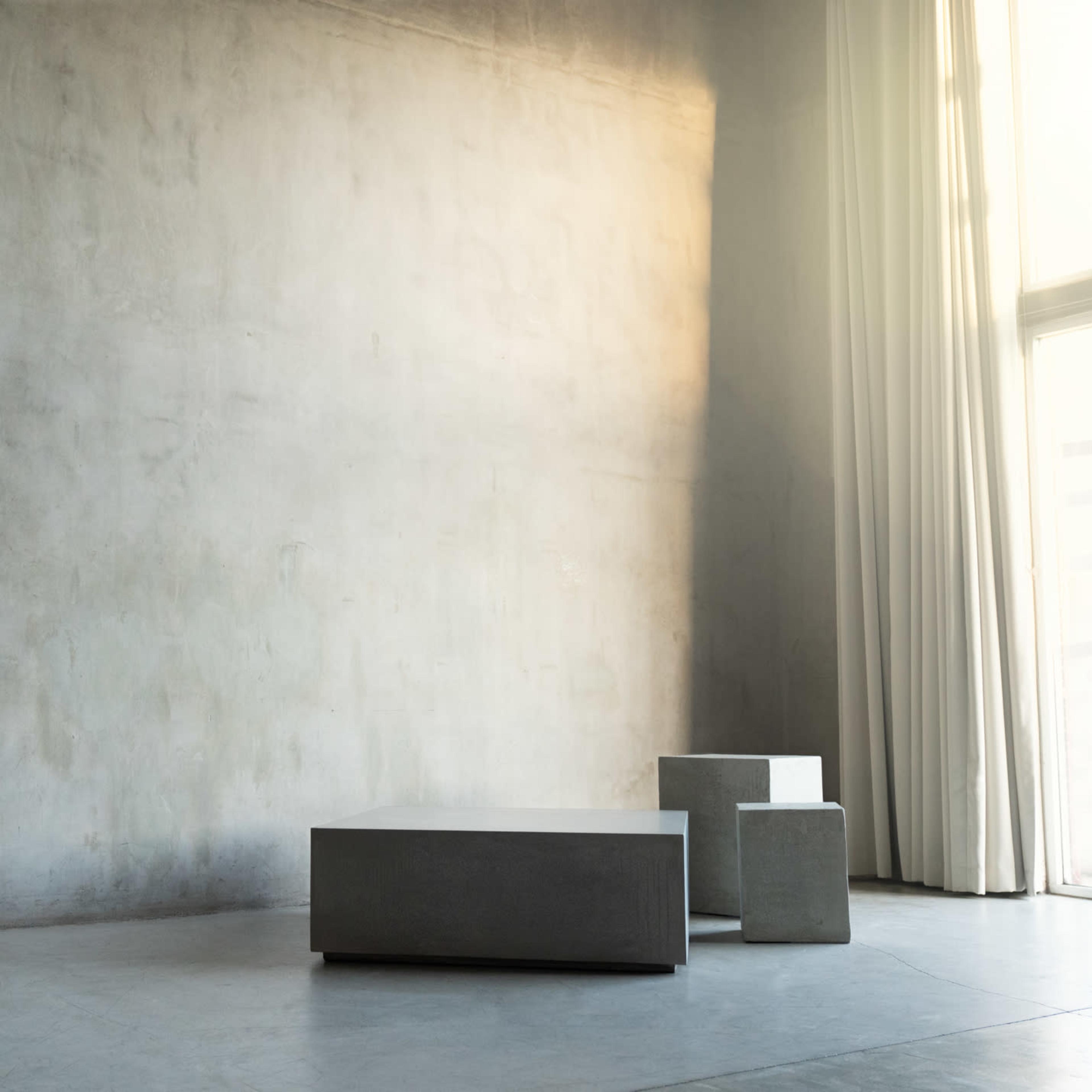 A minimalistic interior space with a textured gray wall, two geometric concrete tables, and large windows covered by sheer curtains.