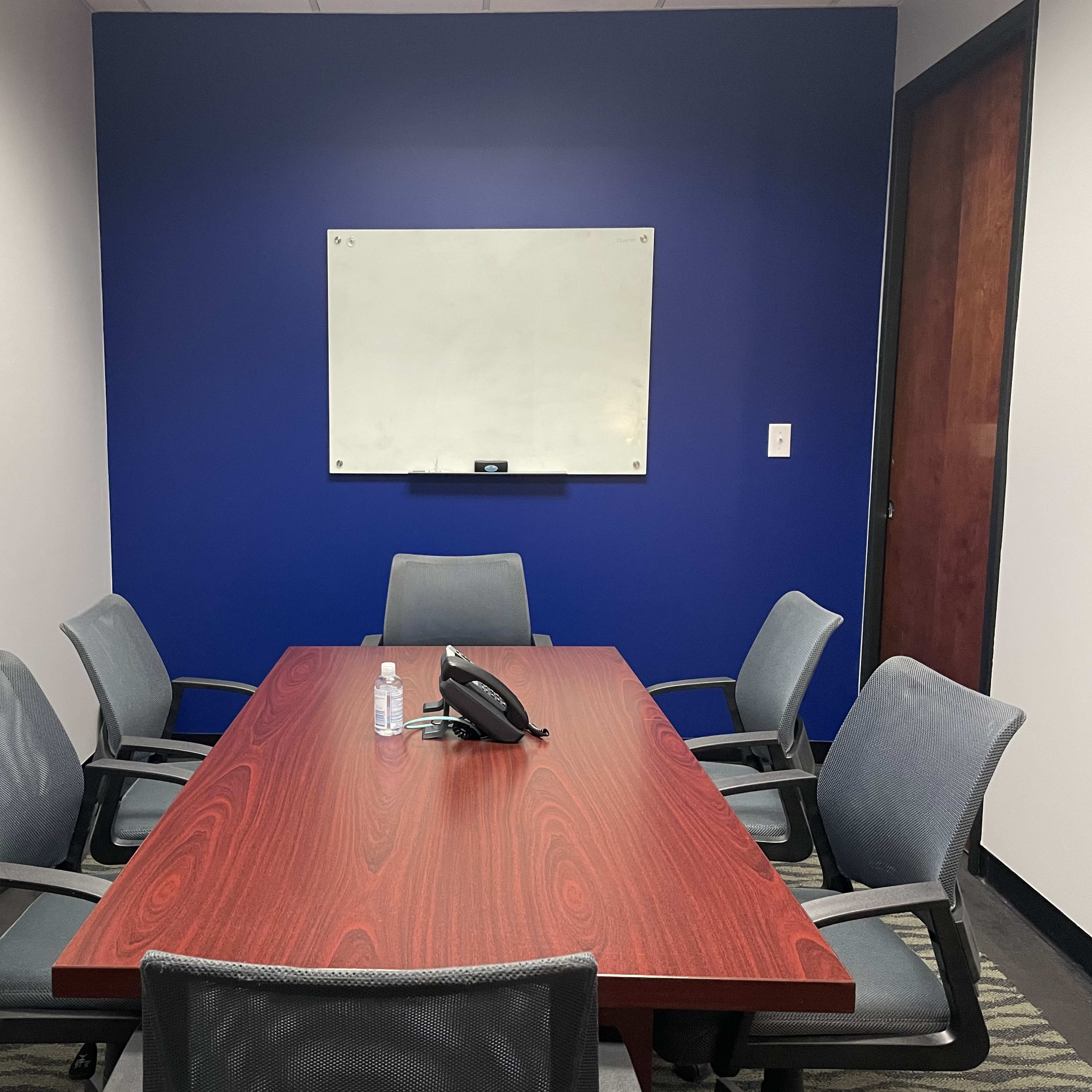 A meeting room with a wooden table surrounded by six gray chairs, a phone in the center, and a whiteboard on the blue wall.