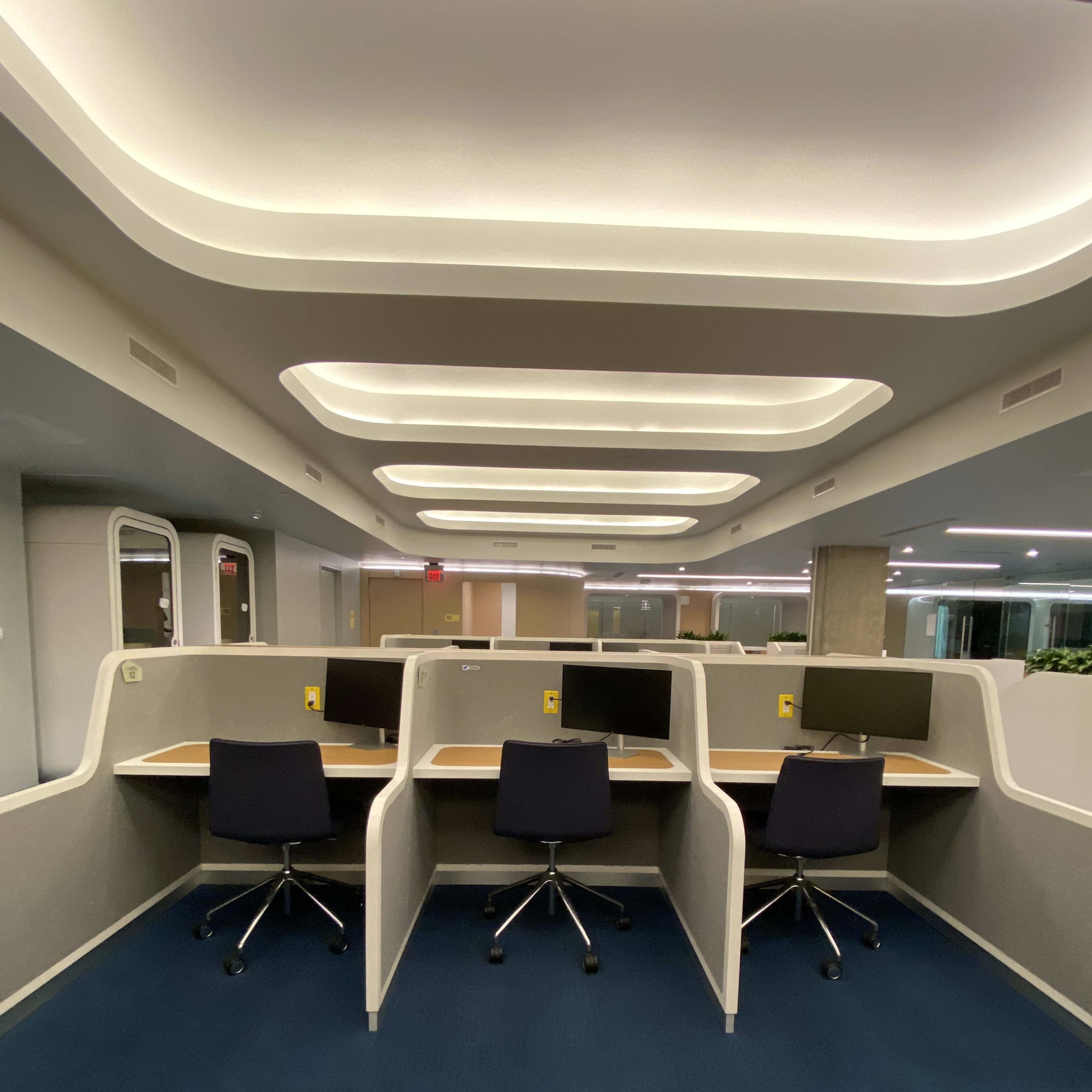 A row of four office workstations with computers, each enclosed by low partitions, situated in a well-lit, modern office space.