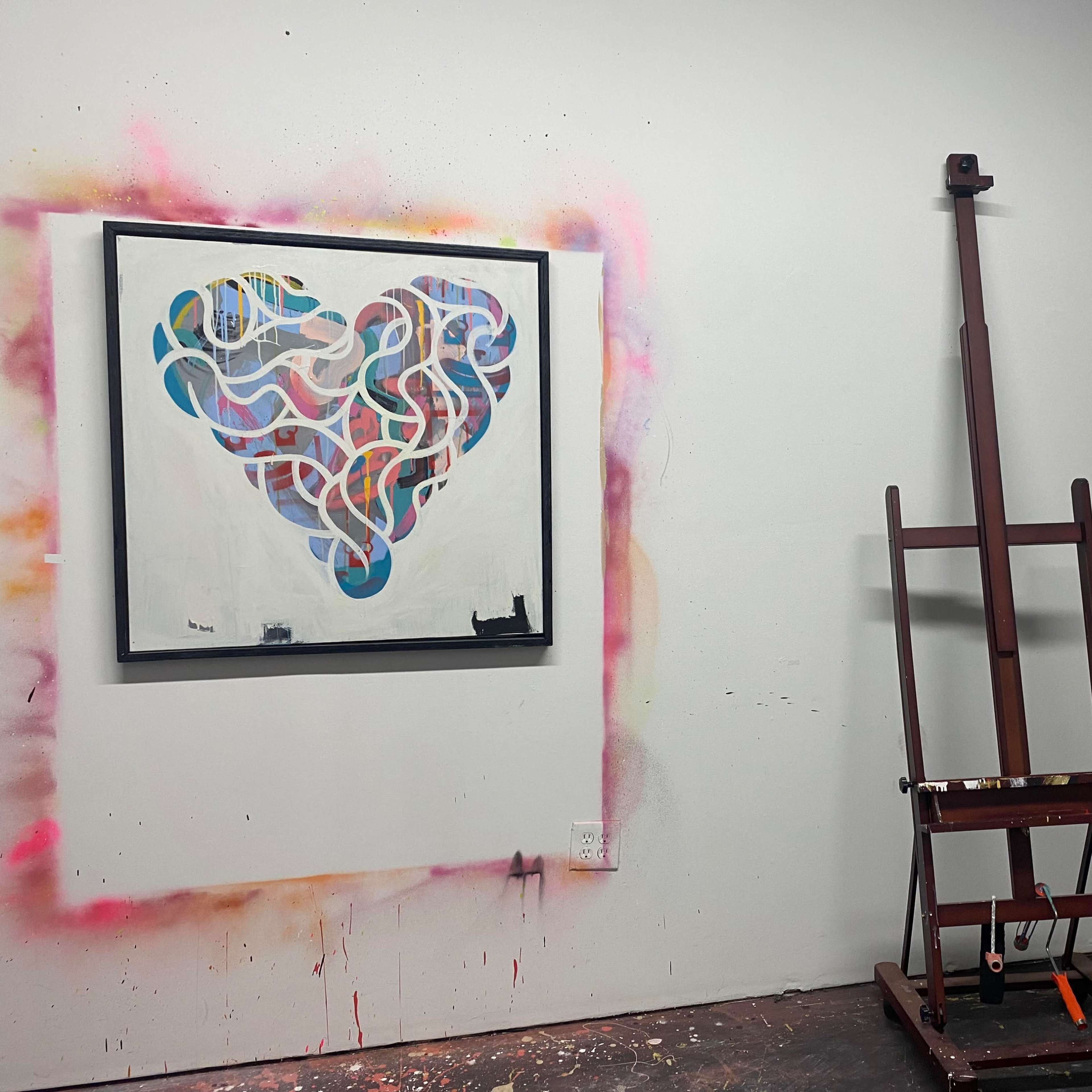 A colorful abstract heart painting on the wall next to a wooden easel in an art studio.