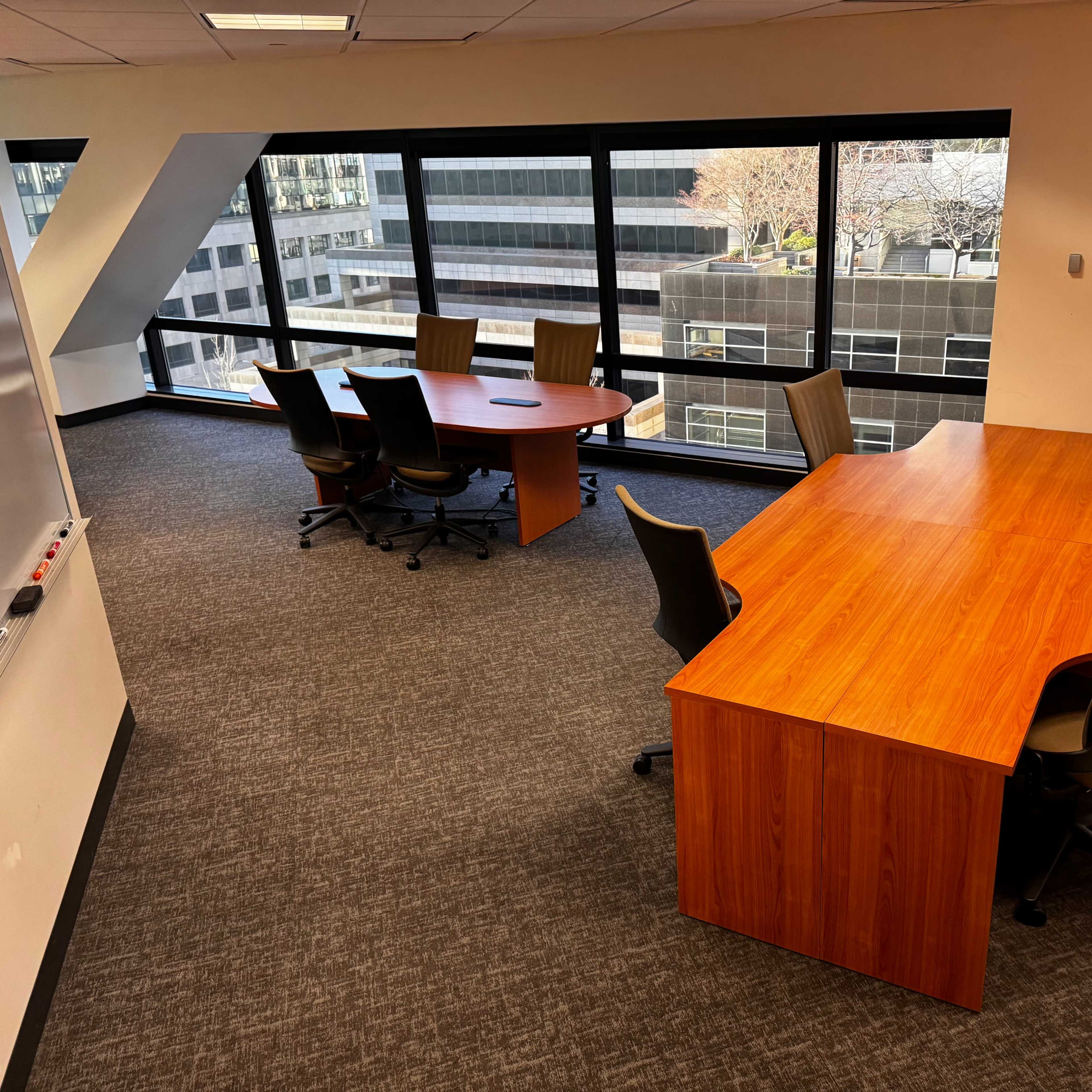 A modern office space featuring two wooden meeting tables surrounded by swivel chairs, with large windows overlooking city buildings.