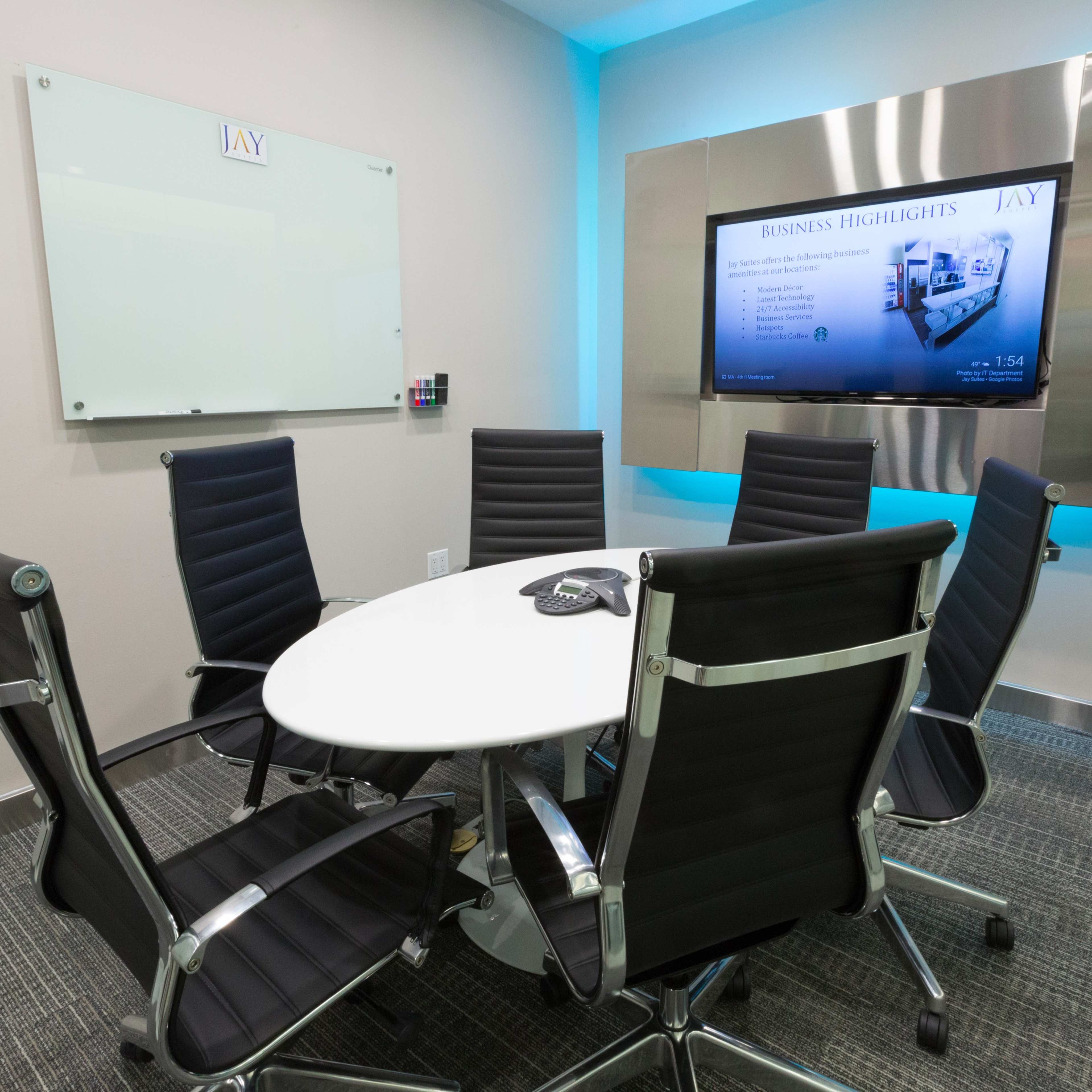 A modern conference room features a round table surrounded by six black chairs, with a large screen displaying business highlights on the wall.