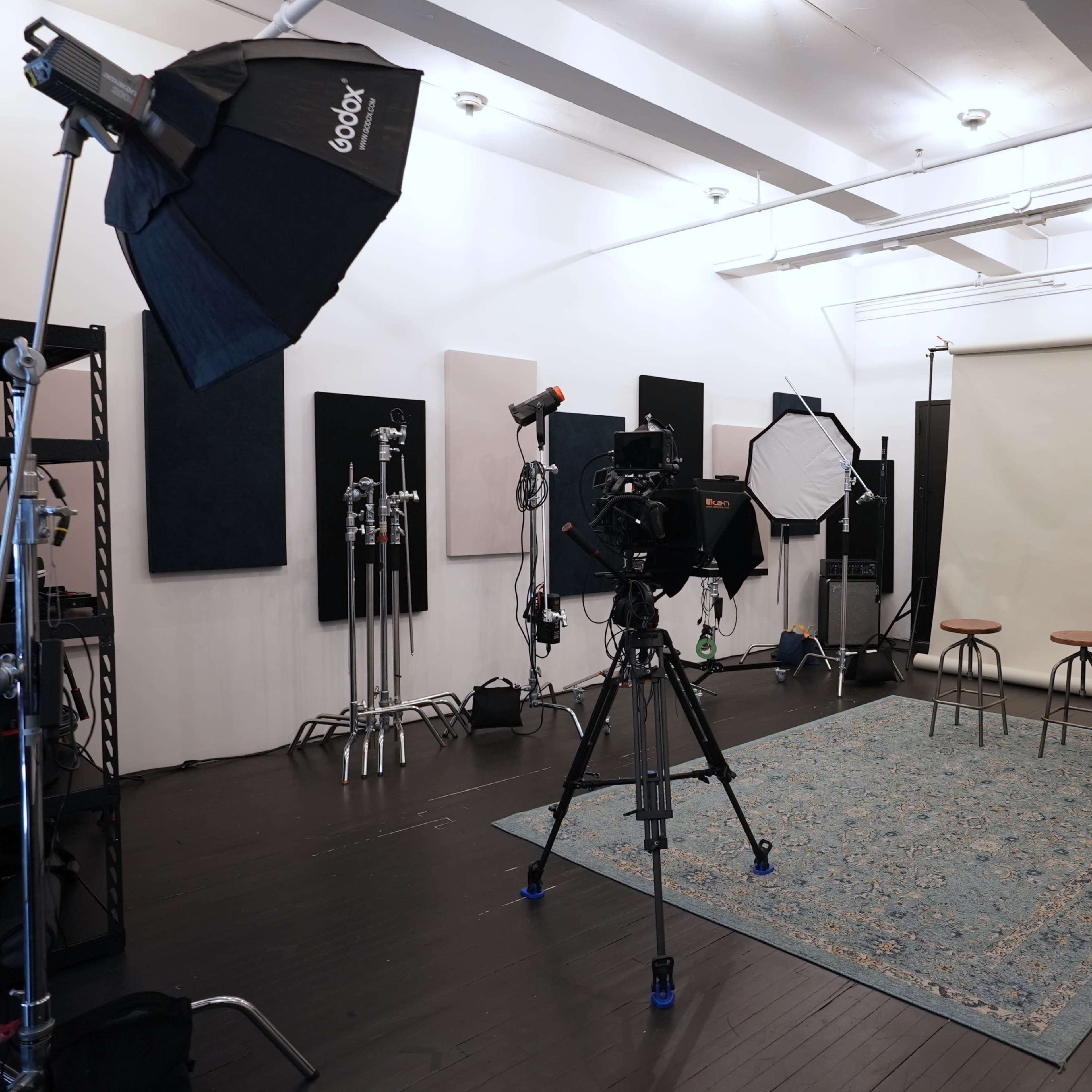 A photography studio is set up with multiple lights, cameras, and two stools against a plain backdrop on a patterned rug.