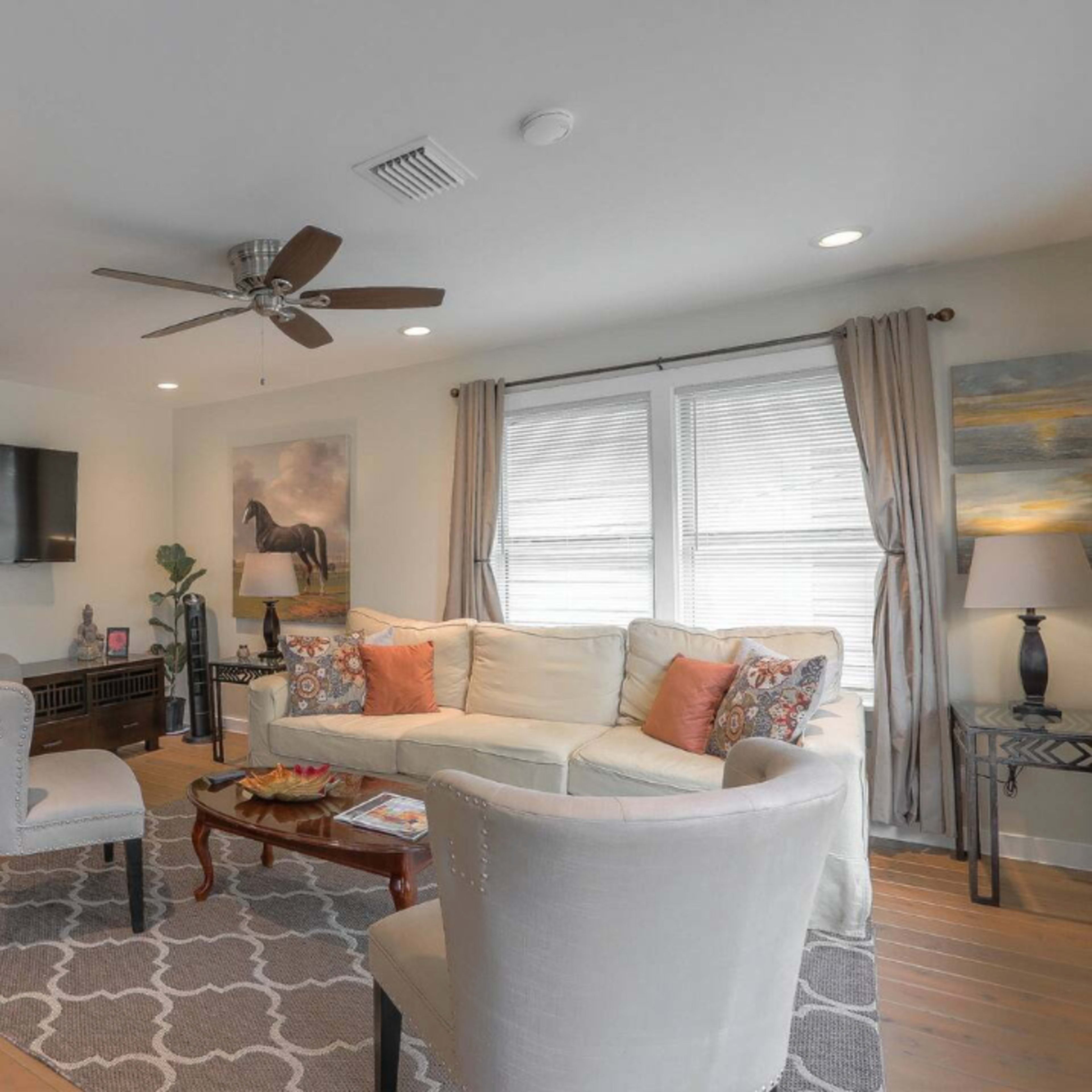 A living room features a white sofa, two light-colored chairs, and a coffee table, with a large window and a television mounted on the wall.