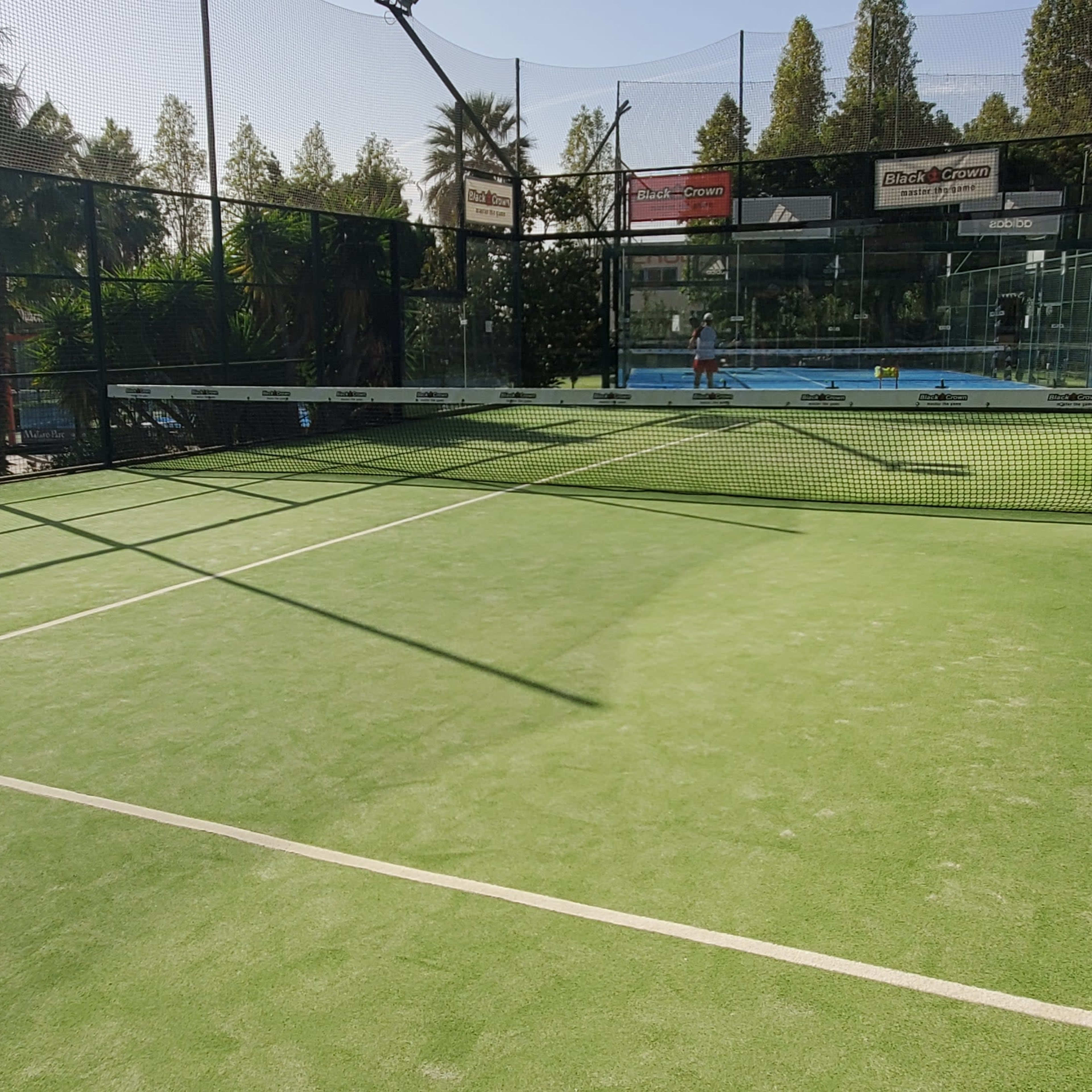 A brightly lit padel court with green artificial turf, surrounded by high fences and palm trees in the background.