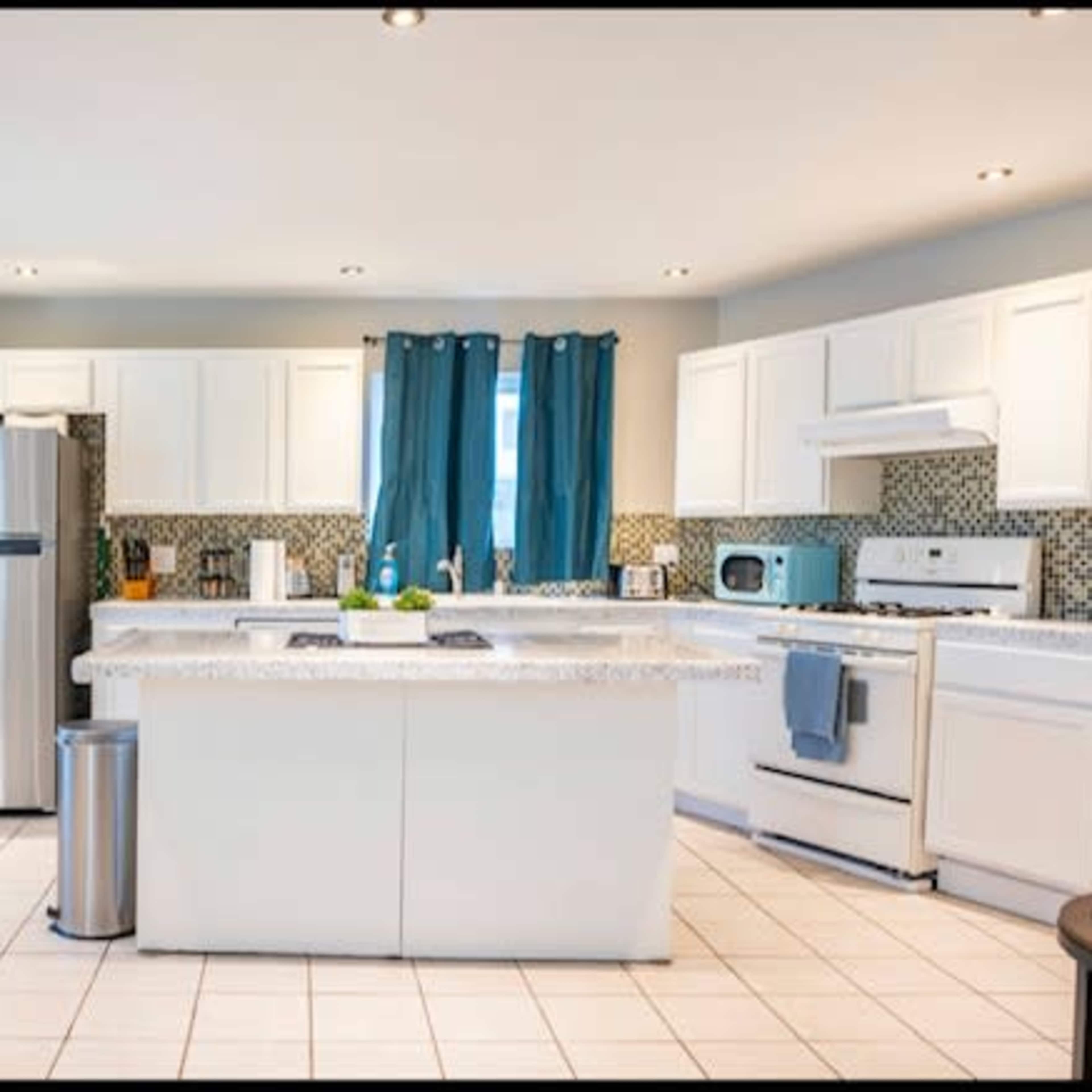 A modern kitchen featuring white cabinets, a large island with a countertop, stainless steel appliances, and blue curtains.