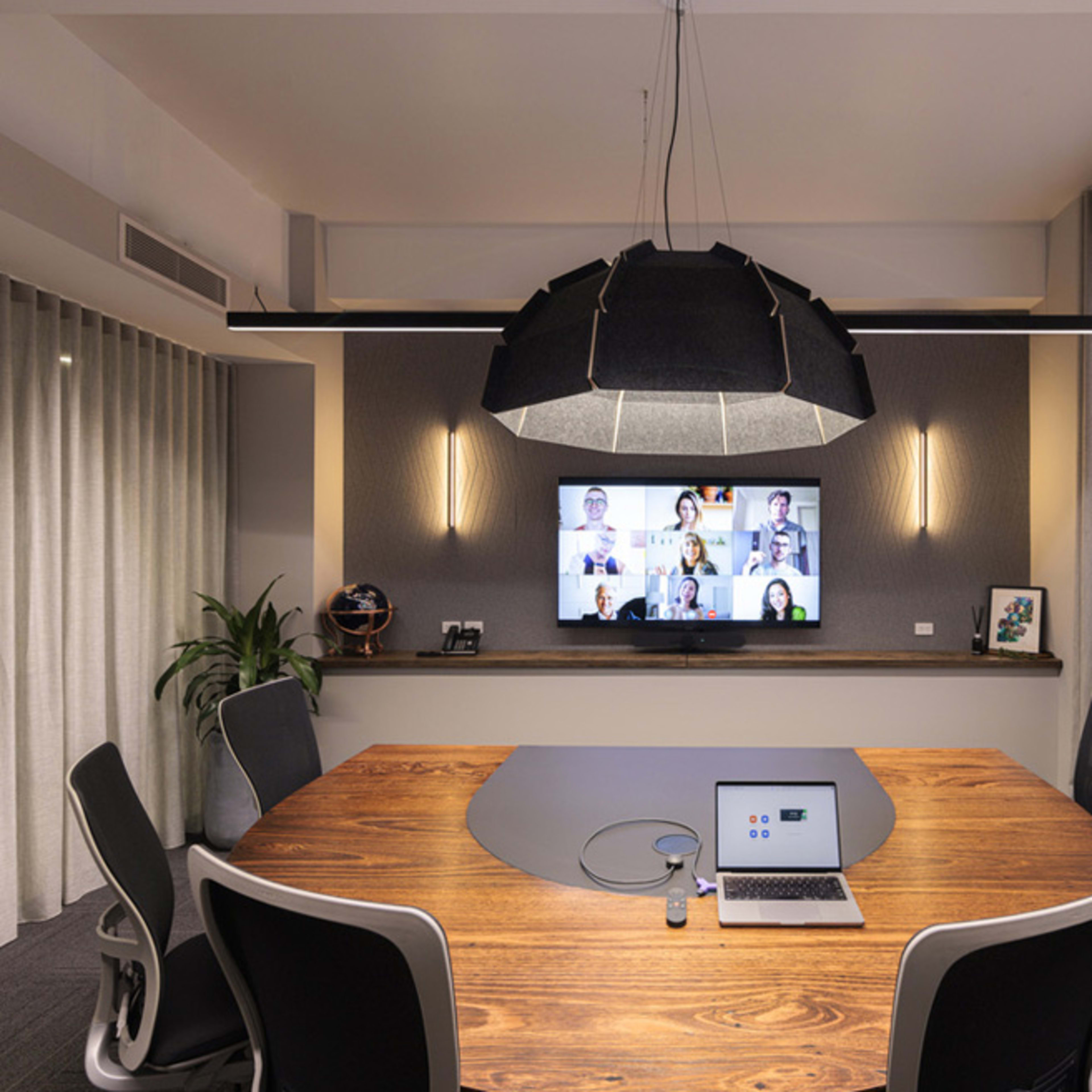 A modern conference room features a large wooden table at the center, surrounded by ergonomic chairs, with a screen on the wall displaying a video call.