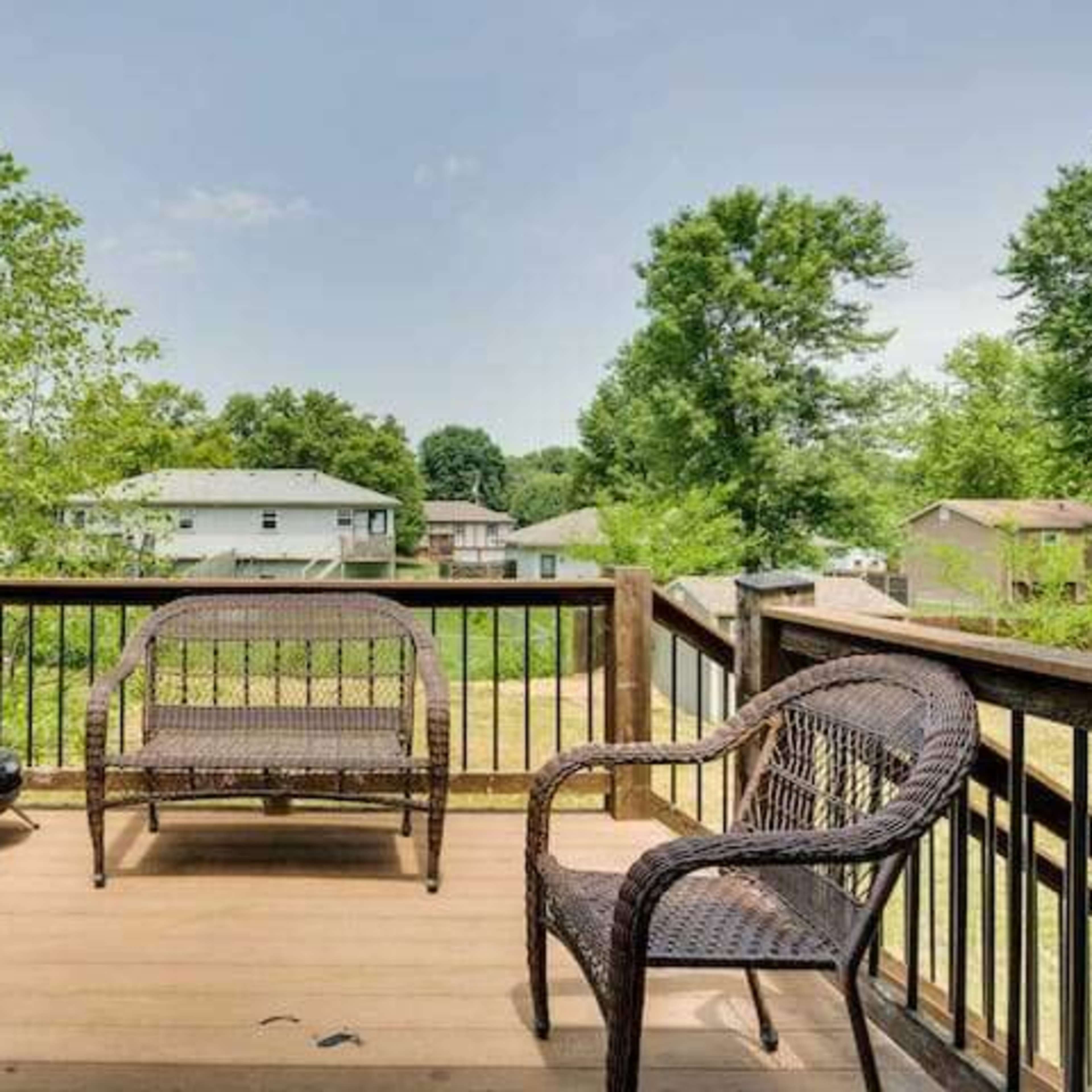 A wooden deck features three wicker chairs, overlooking a grassy yard surrounded by trees and neighboring houses.