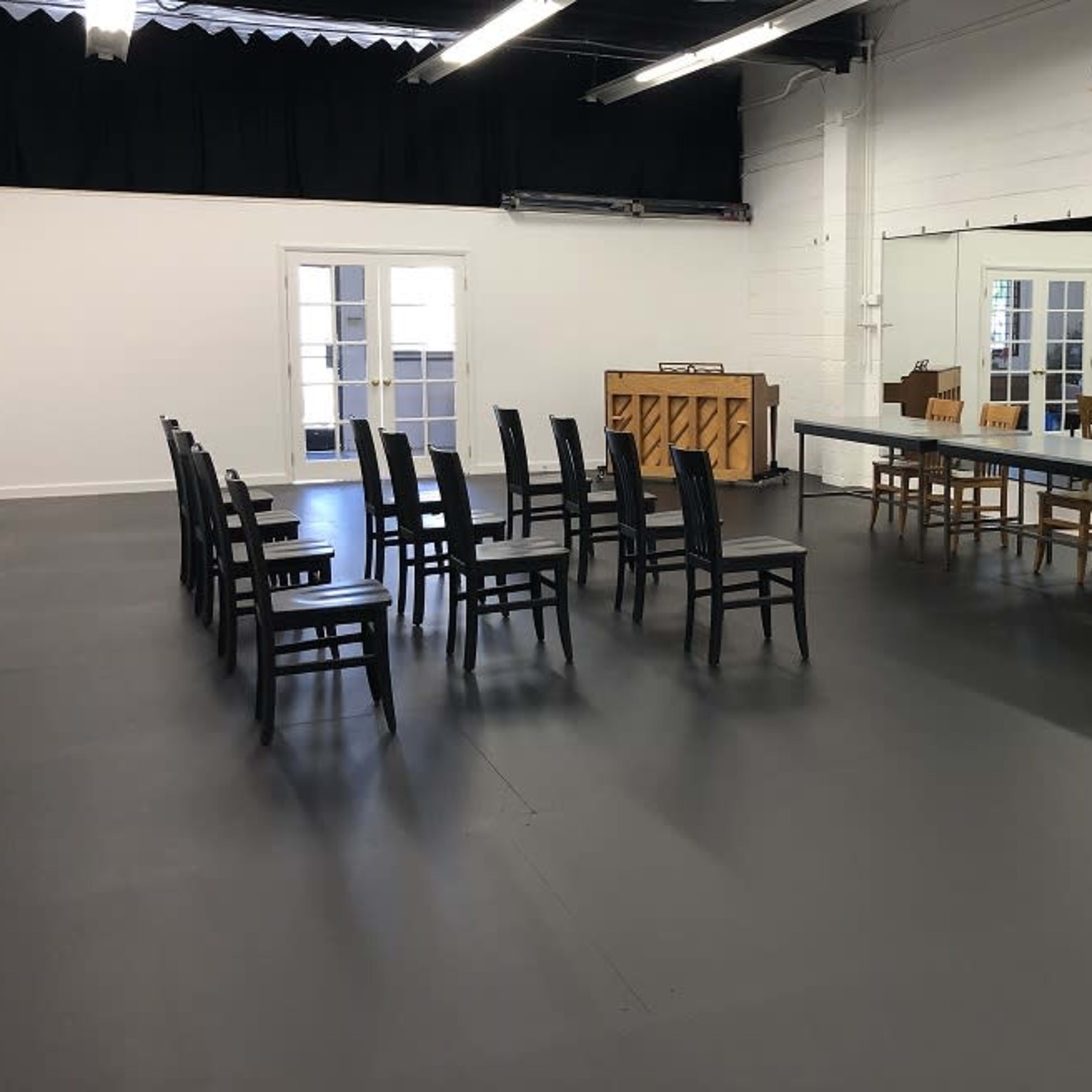 A spacious room with a mirror on one wall, a long table and several wooden chairs arranged in two rows, and a piano in the background.