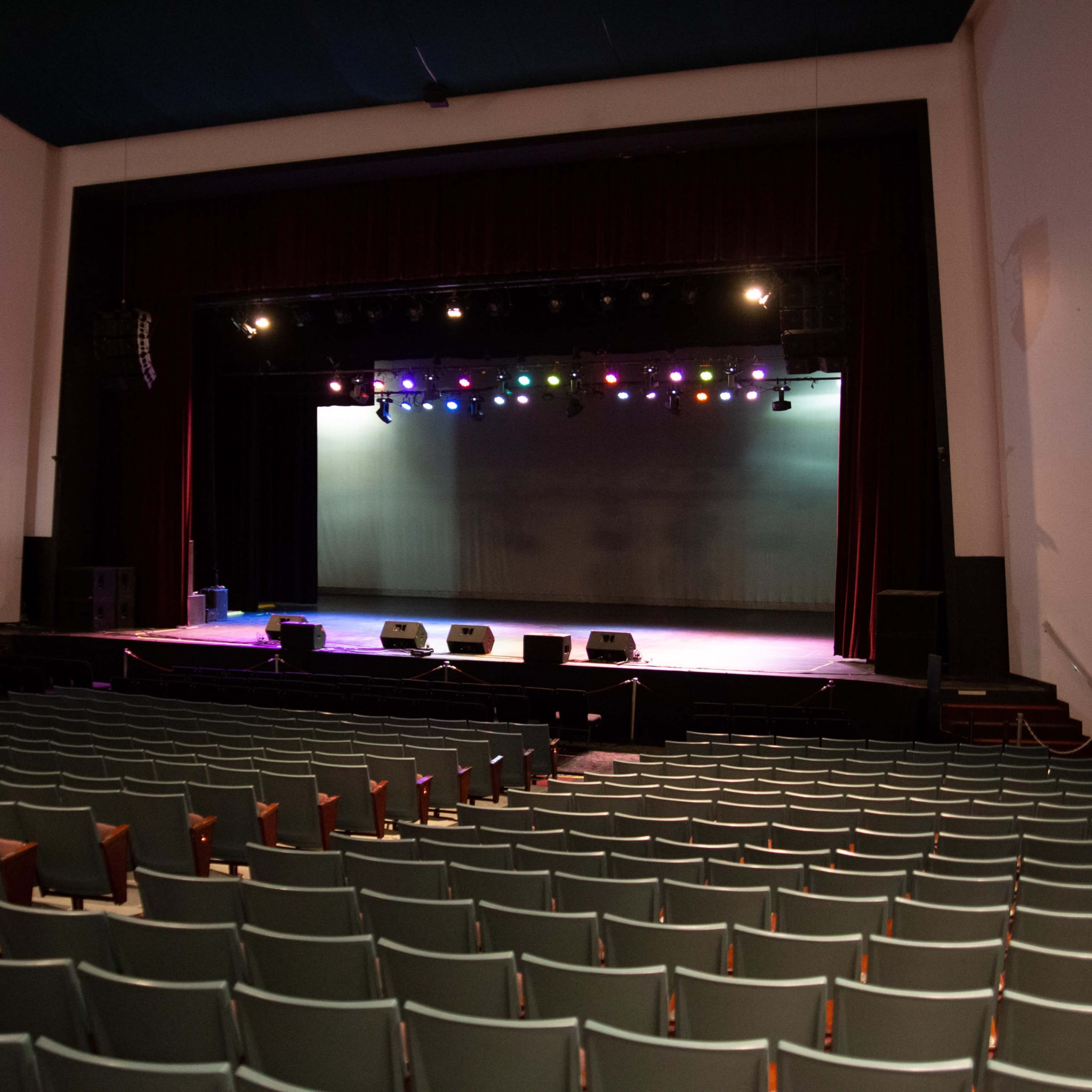 An empty auditorium with rows of seats facing a stage equipped with colorful lights and audio equipment.