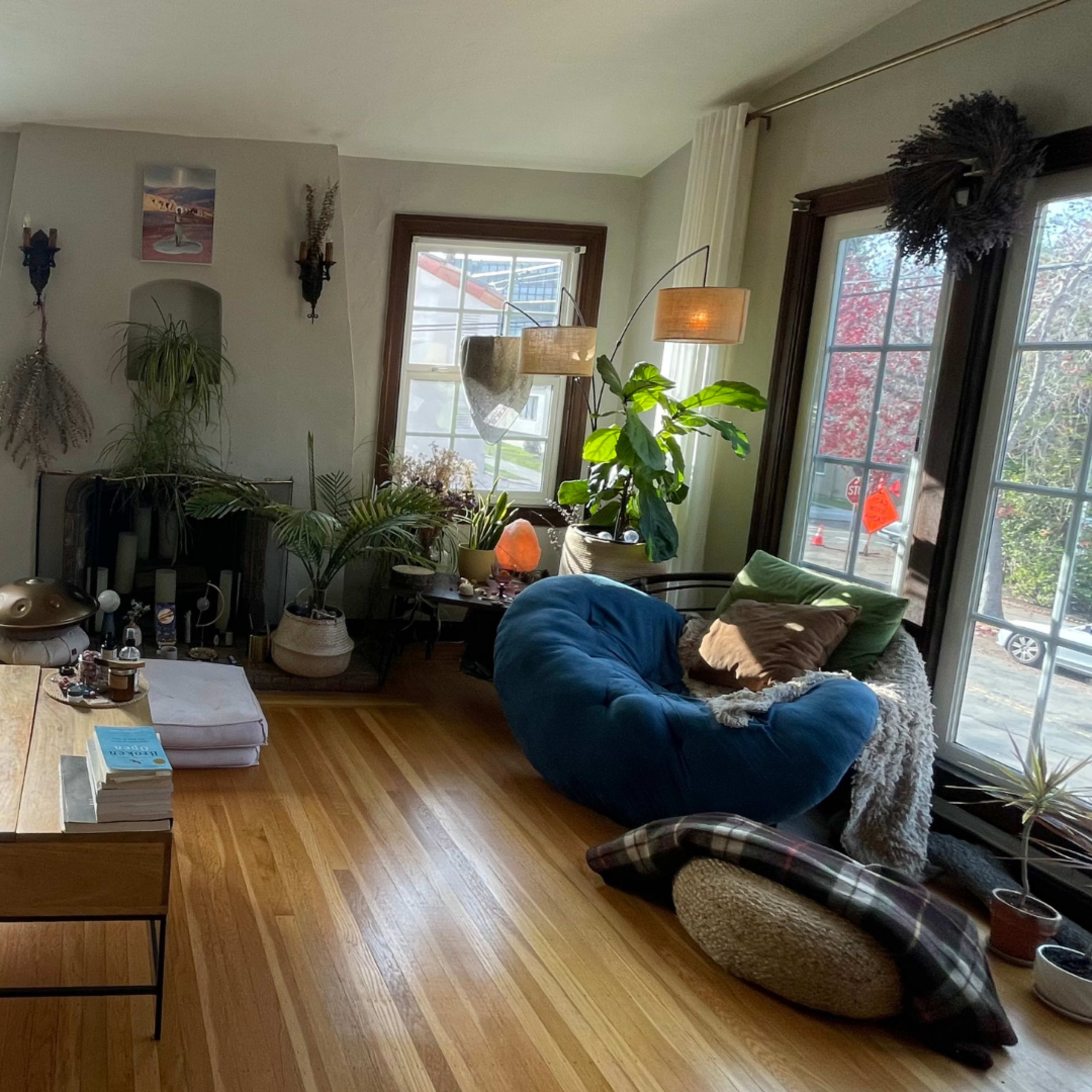 The living room features a green tufted sofa, a wooden coffee table, a beanbag chair, and large windows that provide natural light, along with various plants and decor.