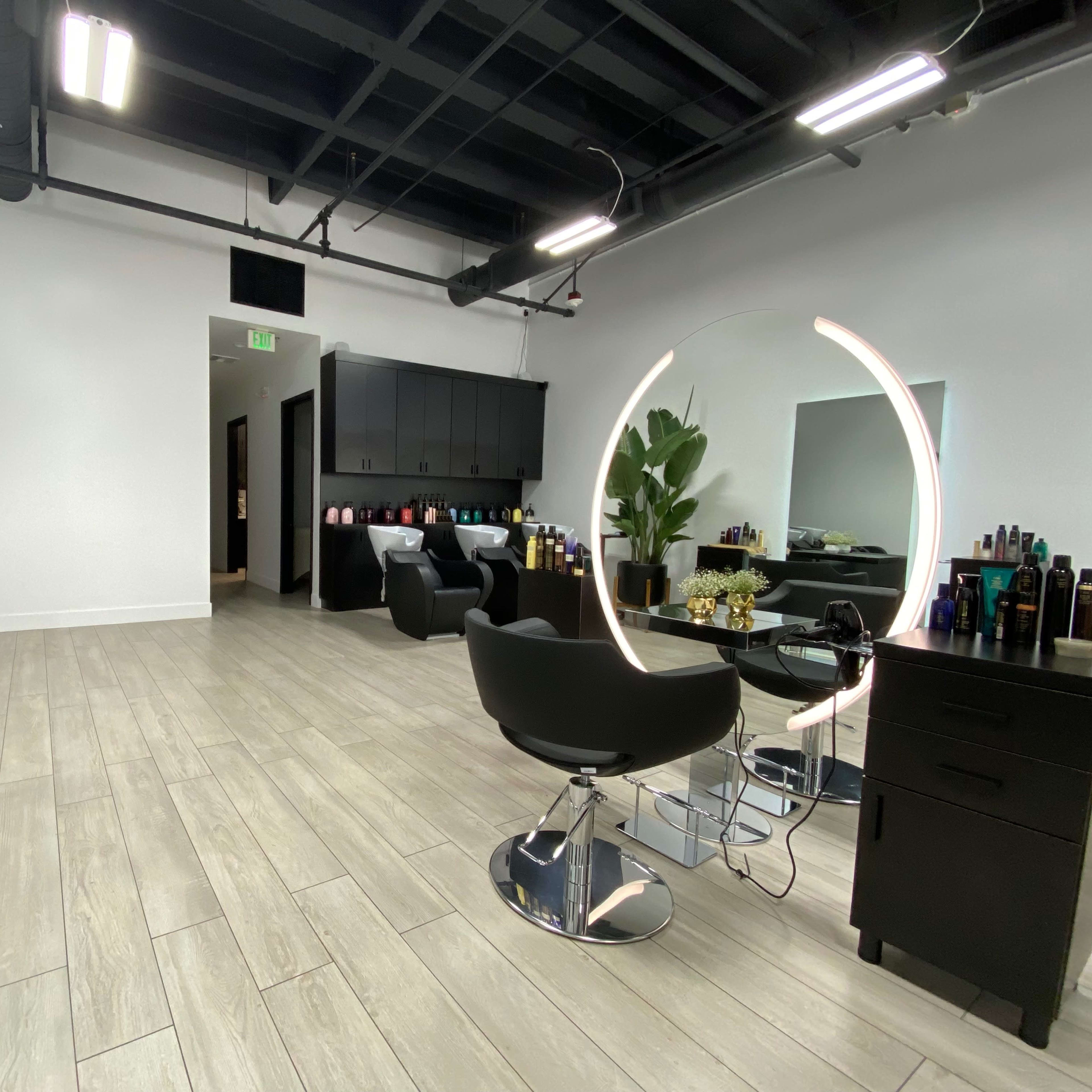 A modern hair salon featuring two stylist chairs with a large illuminated mirror, plants, and a product display along the back wall.