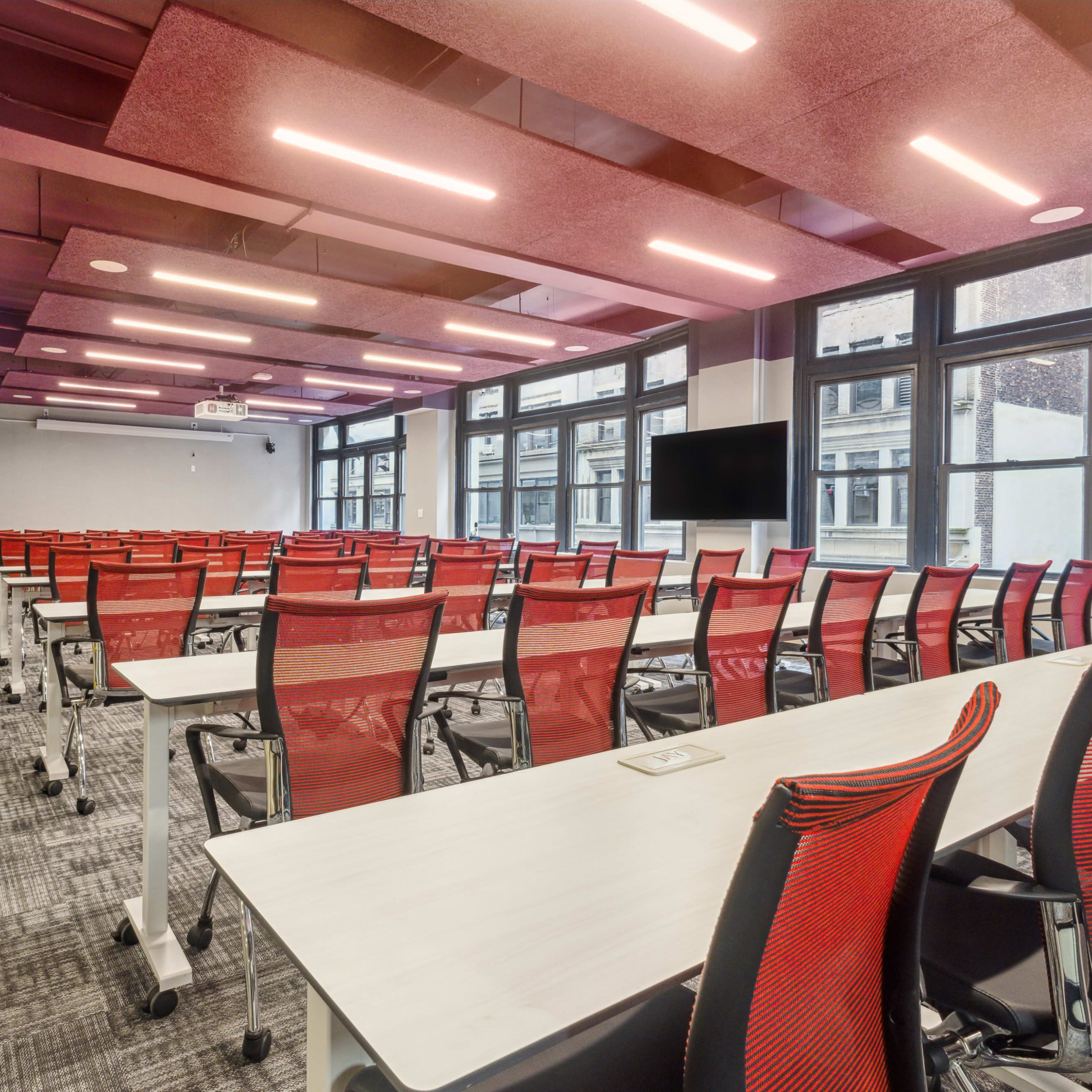 A large classroom features rows of red chair-equipped tables arranged for a presentation, with windows providing ample natural light.
