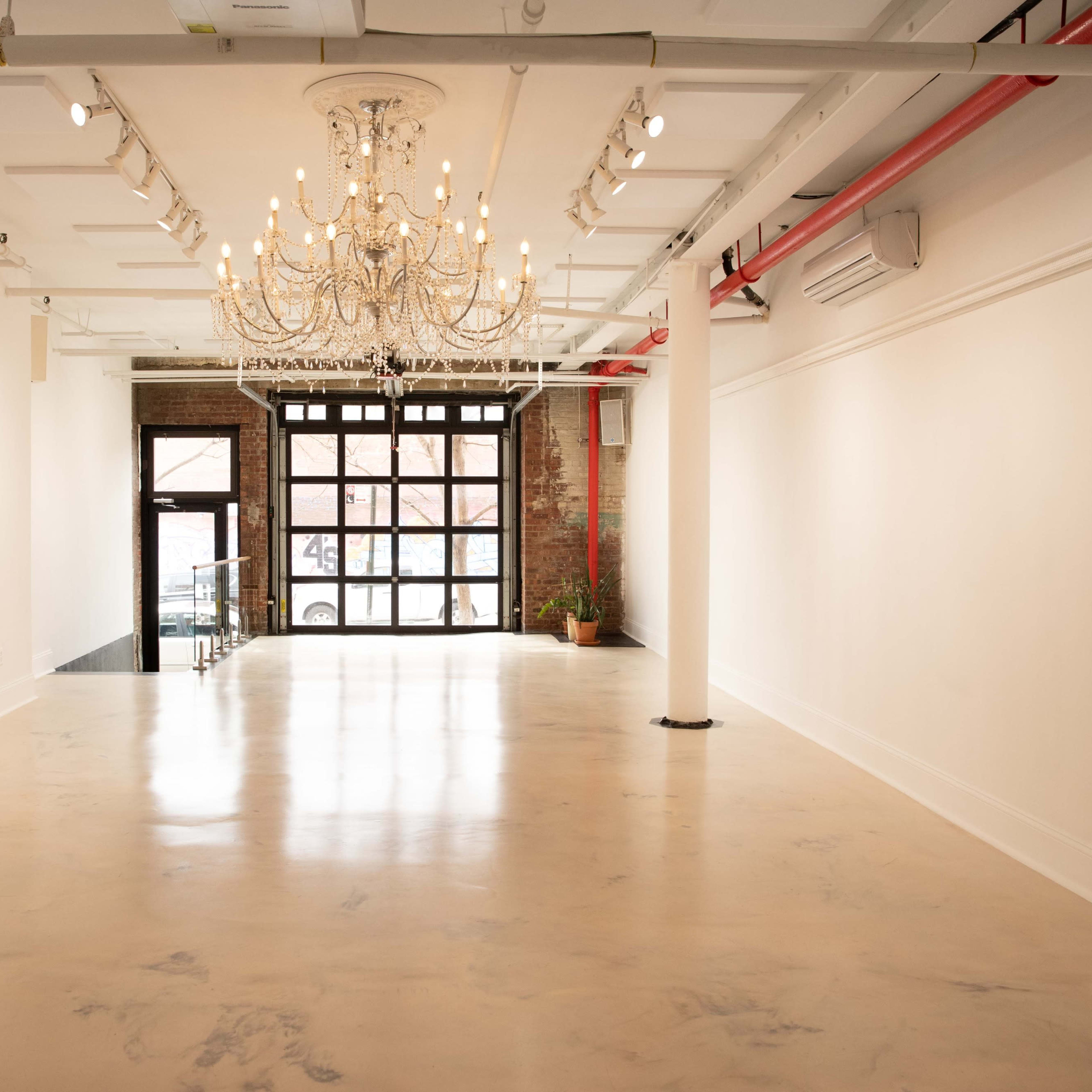 A spacious, empty room with polished concrete flooring, a chandelier hanging from the ceiling, and large windows at the far end.