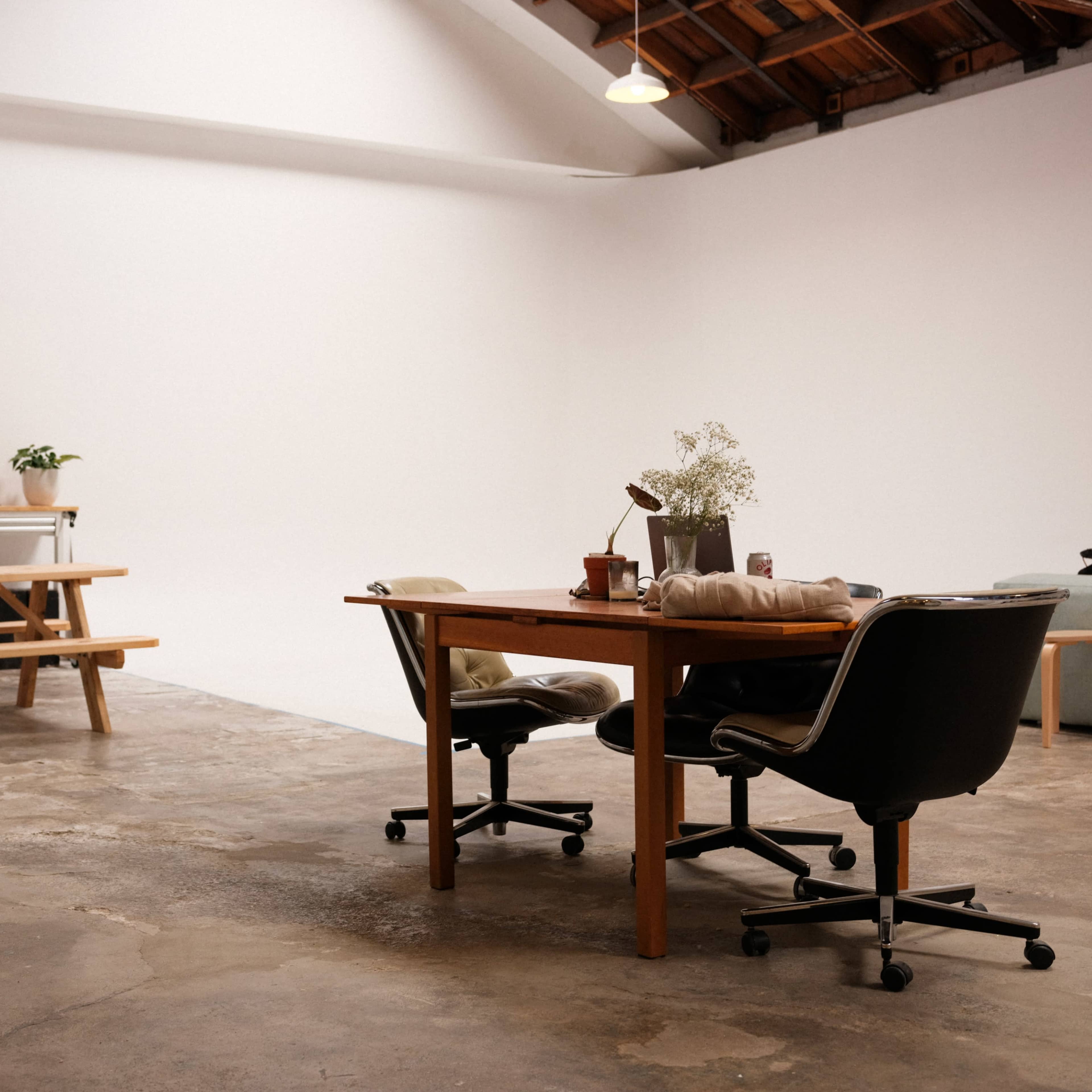 A spacious, minimalist studio featuring a wooden table with chairs, a bench, and a white wall in the background.