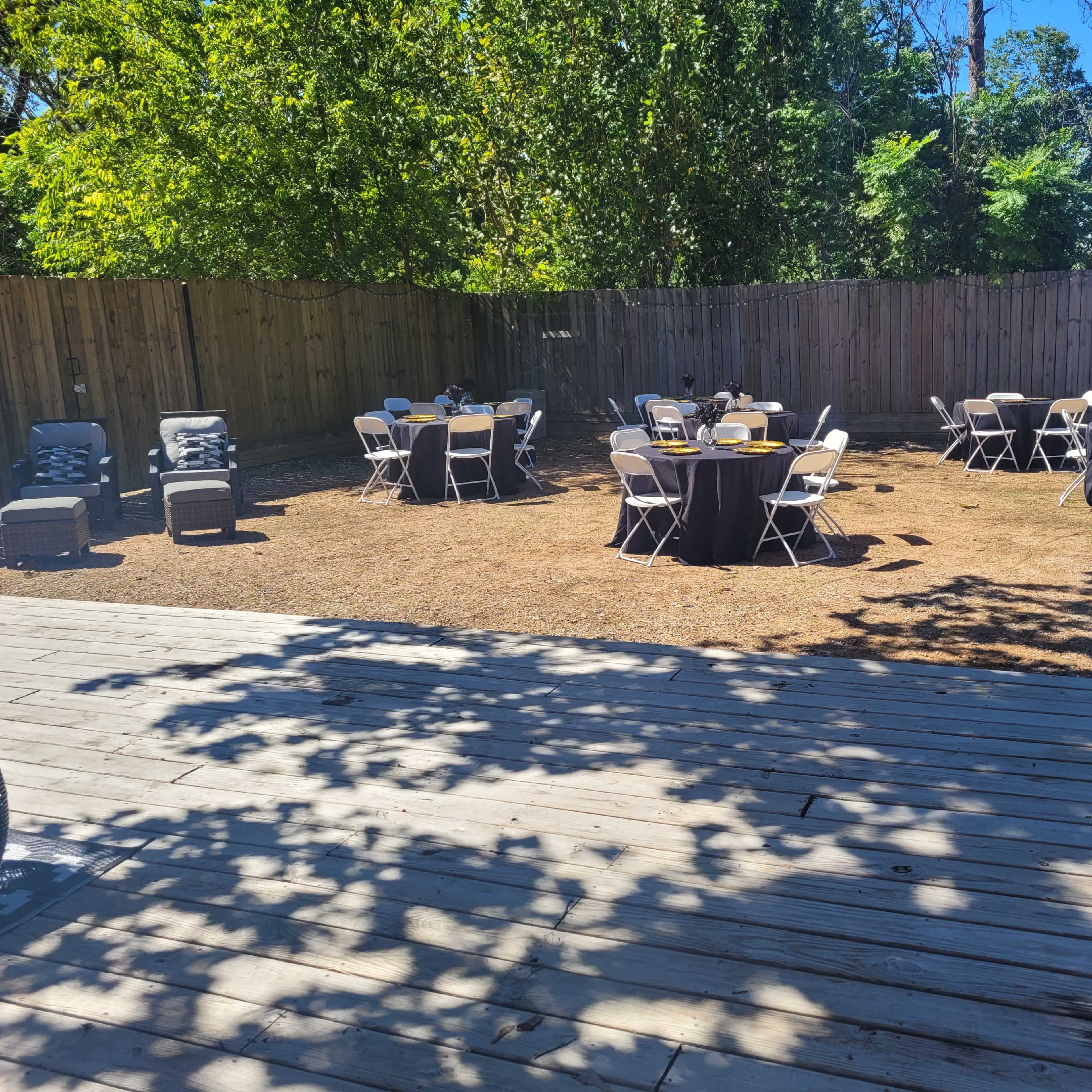 A spacious outdoor area set up for an event, featuring round tables with black tablecloths arranged on grass under trees, and a wooden deck in the foreground.