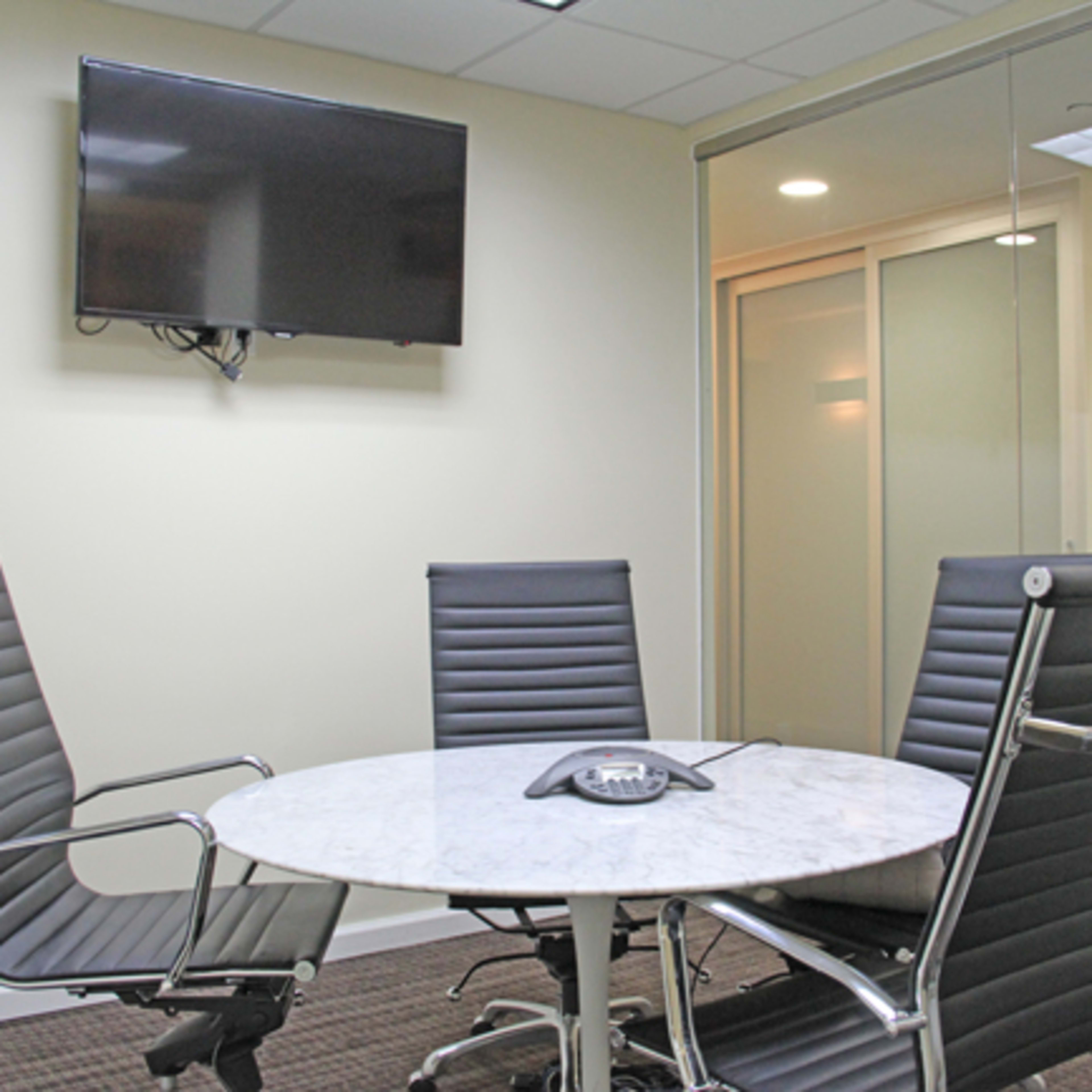 A conference room features a round table with four black leather chairs, a telephone in the center, and a wall-mounted television.