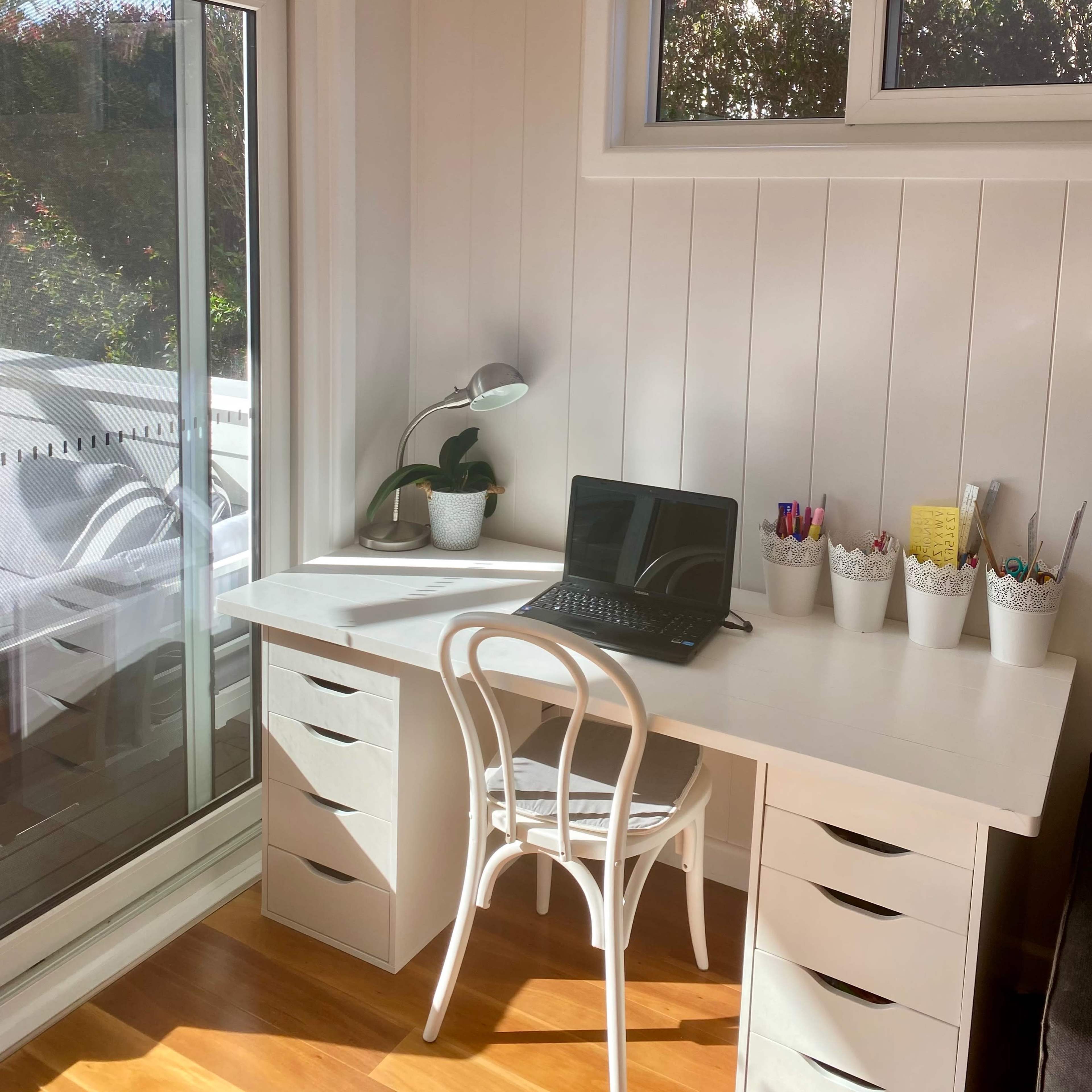 A bright office space features a white desk with a laptop, surrounded by organized containers of pens and a small plant, beside a large window.