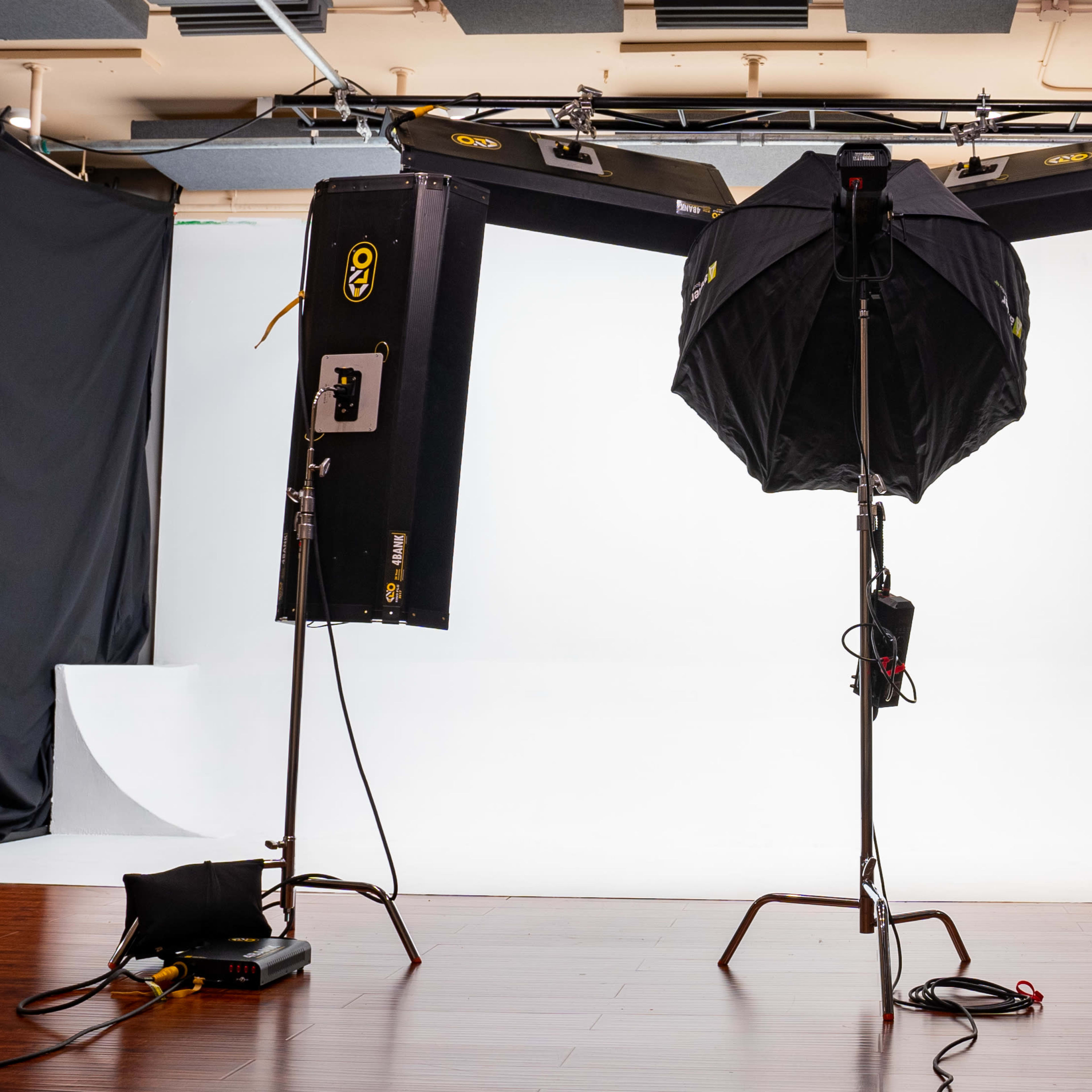 A photography studio setup featuring three lighting equipment stands and a large softbox on a central tripod, all facing a white backdrop.