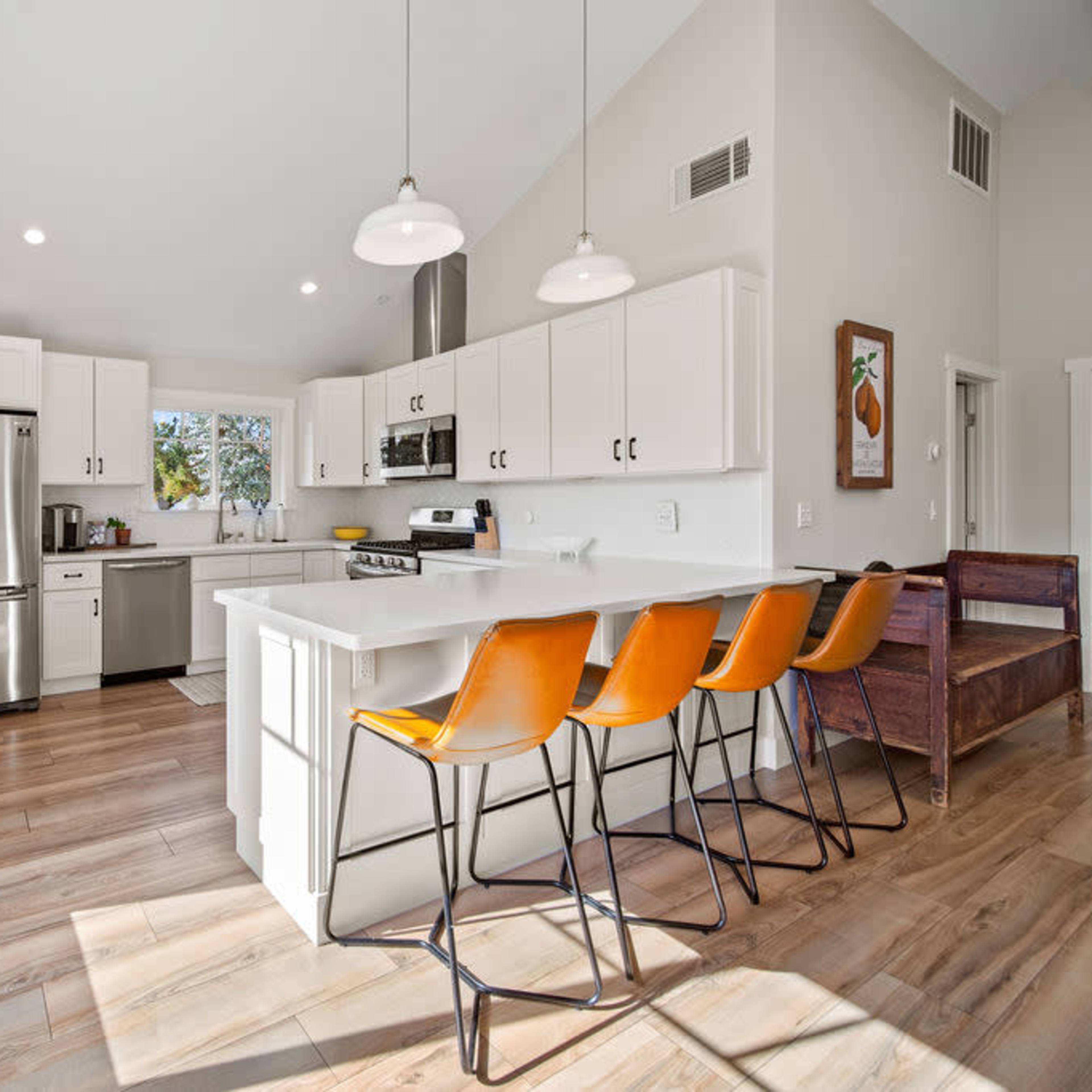 A modern kitchen features white cabinetry, stainless steel appliances, and four orange bar stools at a central island.