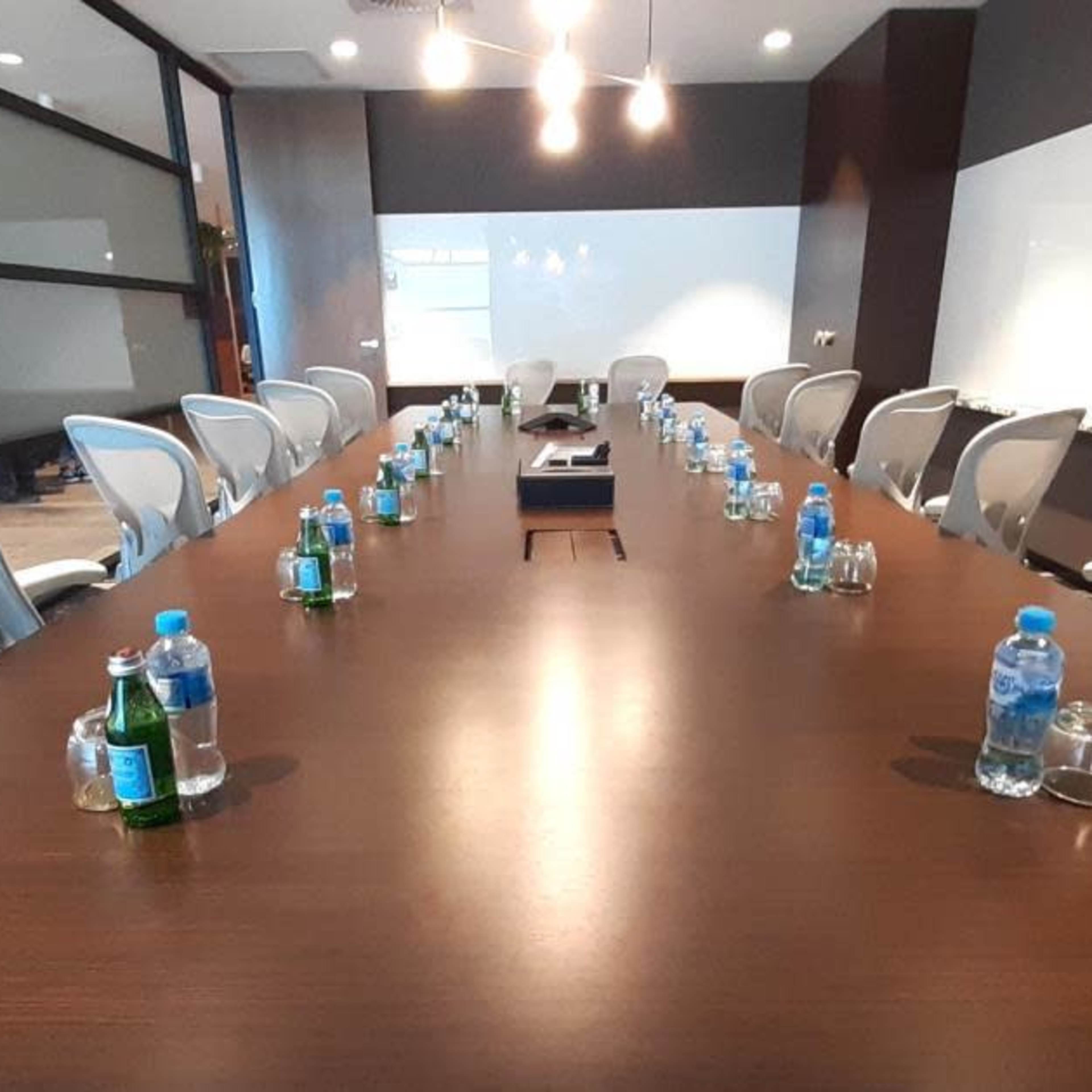 A long, wooden conference table is arranged with water bottles and glasses, surrounded by several chairs in a modern meeting room.