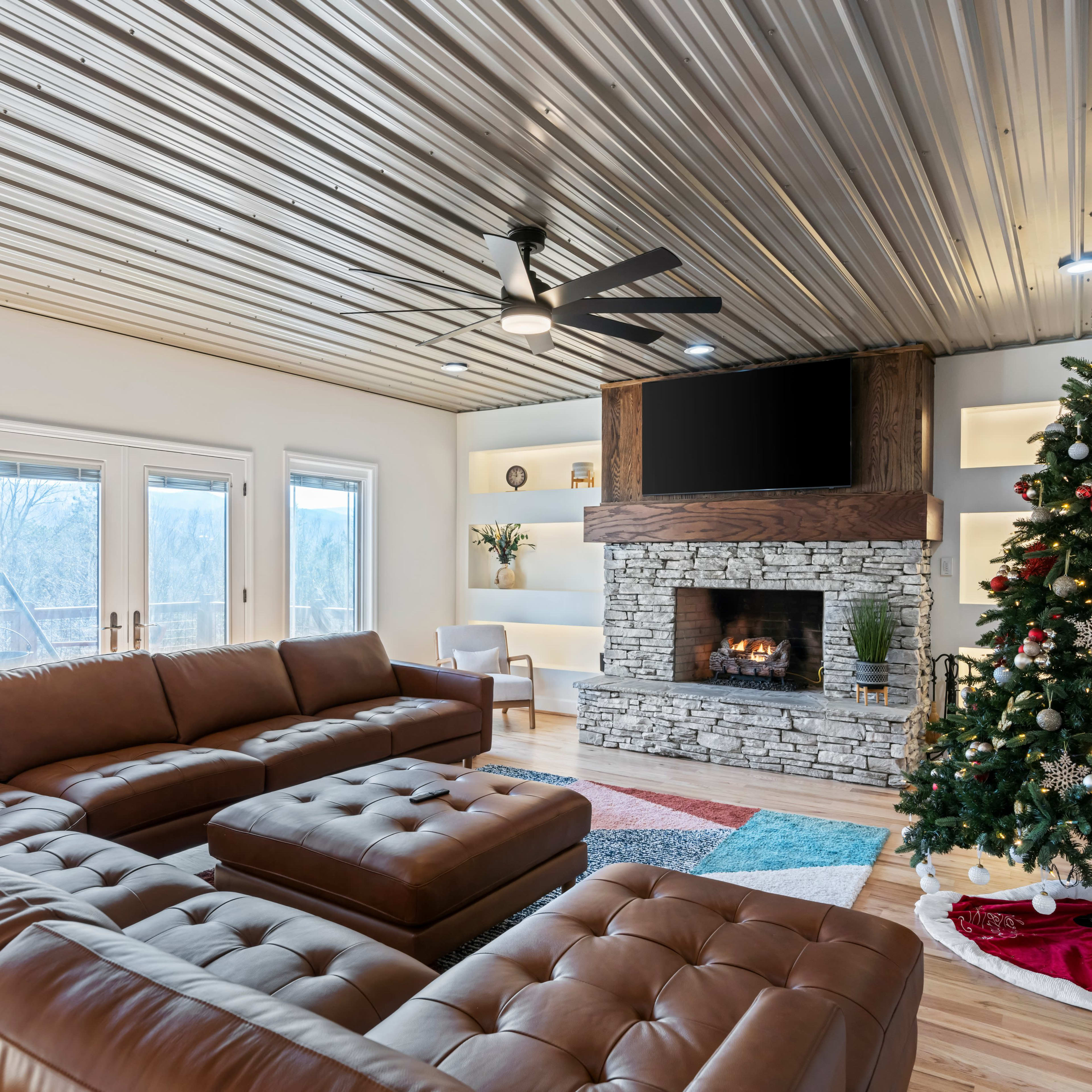 A modern living room features a leather sectional sofa, a stone fireplace with a television above, and a decorated Christmas tree.