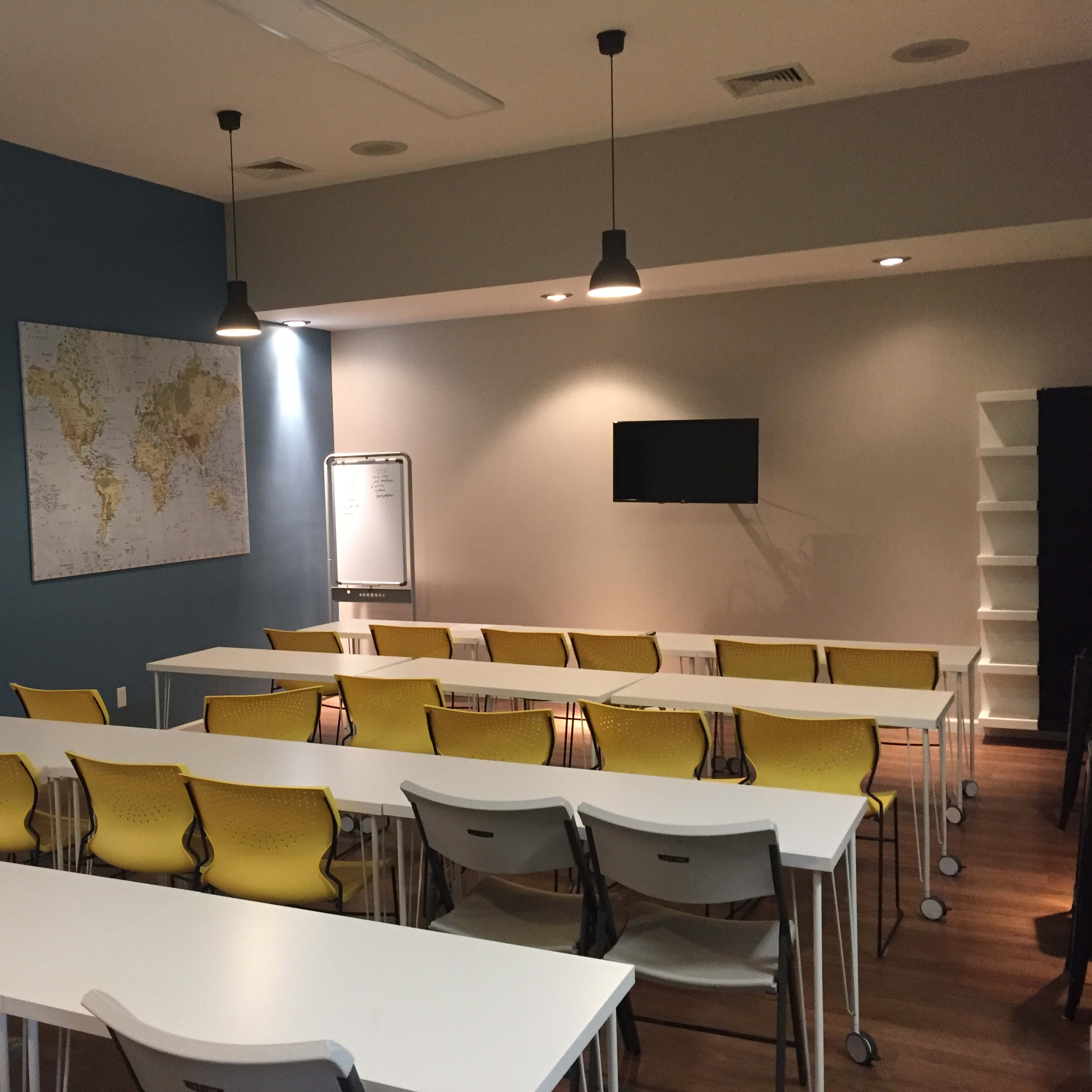 A classroom features yellow chairs and white tables arranged in rows, with a world map on the wall and a television mounted above a whiteboard.